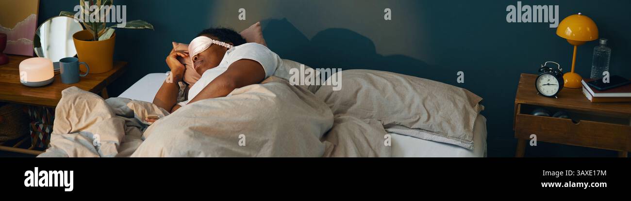 Uomo afro-americano disteso a letto, che dorme tranquillamente in un'accogliente camera da letto. Luci soffuse che creano un'atmosfera tranquilla e rilassata con vari articoli sui letti Foto Stock