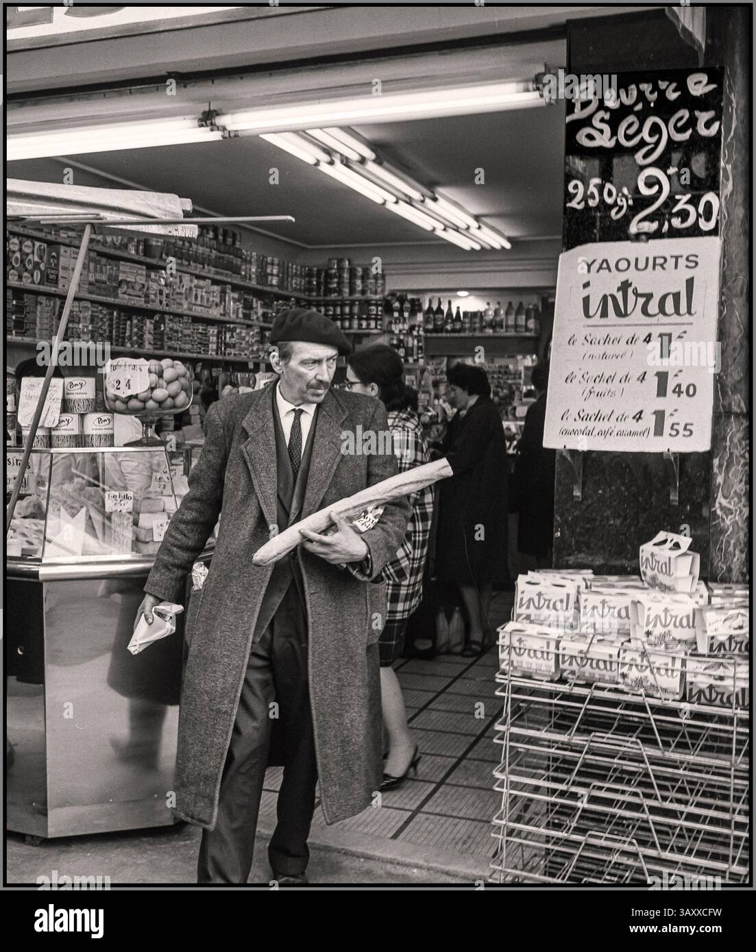 Stile di vita retrò vintage Parigi 1965 con un tipico uomo d'affari francese in fretta, che indossa un berretto che lascia un negozio di gastronomia con provviste tra cui una tipica baguette francese Parigi Francia: Willem Van de poll 1965 Parigi Francia Foto Stock
