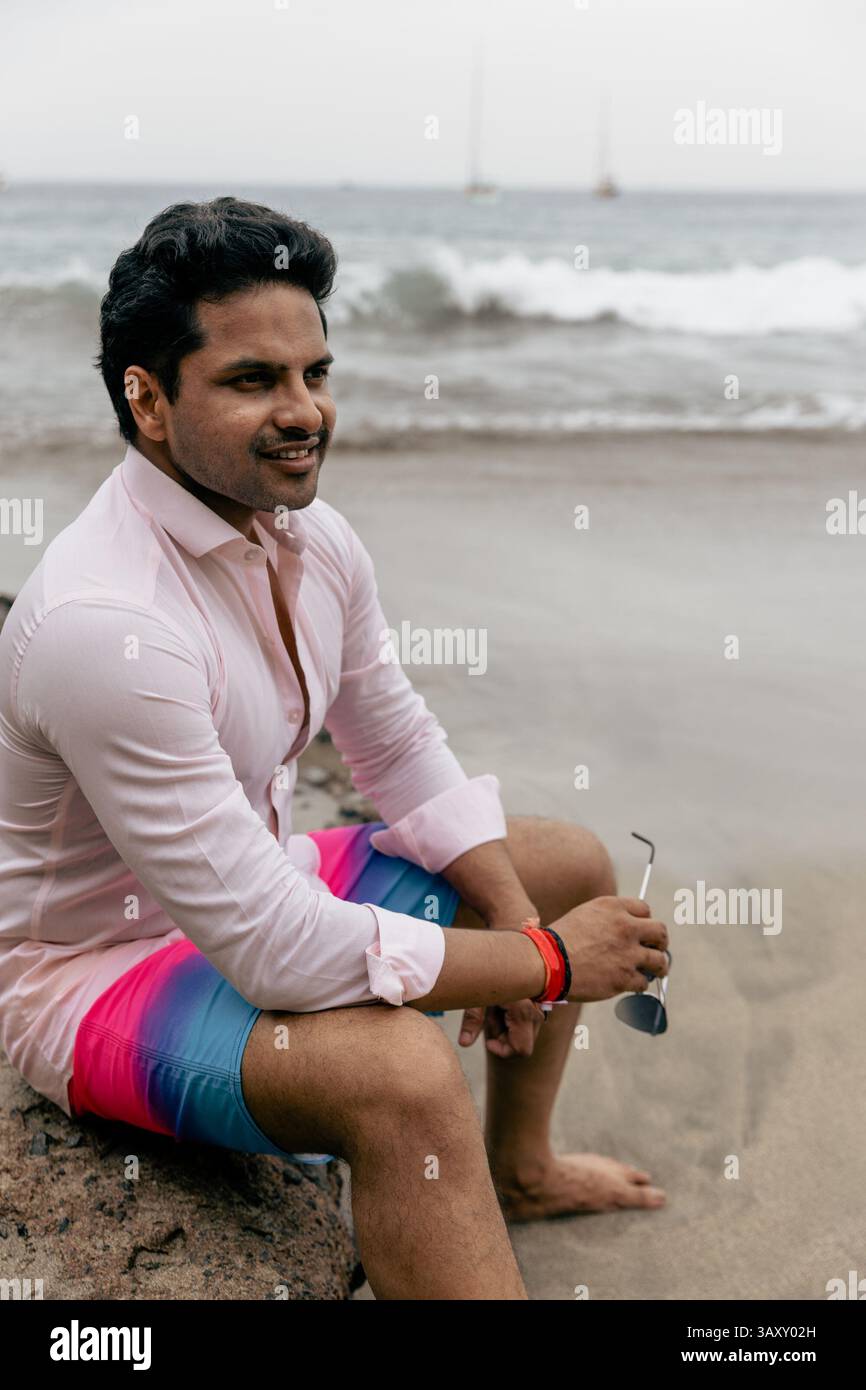 Un uomo è seduto su una roccia su una spiaggia e indossa una camicia rosa e pantaloncini blu e rosa. Sta tenendo gli occhiali da sole Foto Stock
