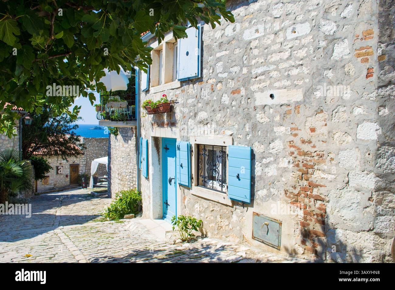Una tranquilla strada acciottolata si snoda attraverso vecchie mura di pietra e persiane blu in Italia, dove la storia permane al sole e all'ombra della vita quotidiana. Foto Stock