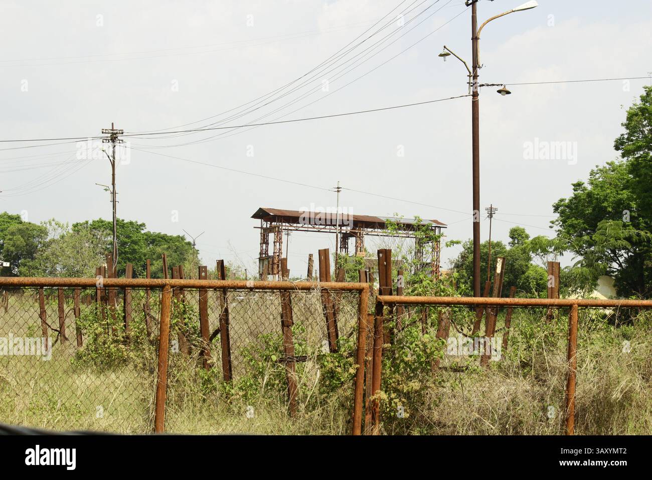 Sito industriale abbandonato con recinzioni arrugginite e vegetazione sovrastata, circondato da linee elettriche e resti di vecchie infrastrutture Foto Stock