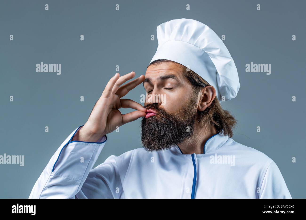 Lo chef fornello in grembiule mostra segni di buon gusto, gesto bellissimo, bacio dita sfondo grigio isolato. Ritratto di un cuoco felice. Cappello da cucina Foto Stock