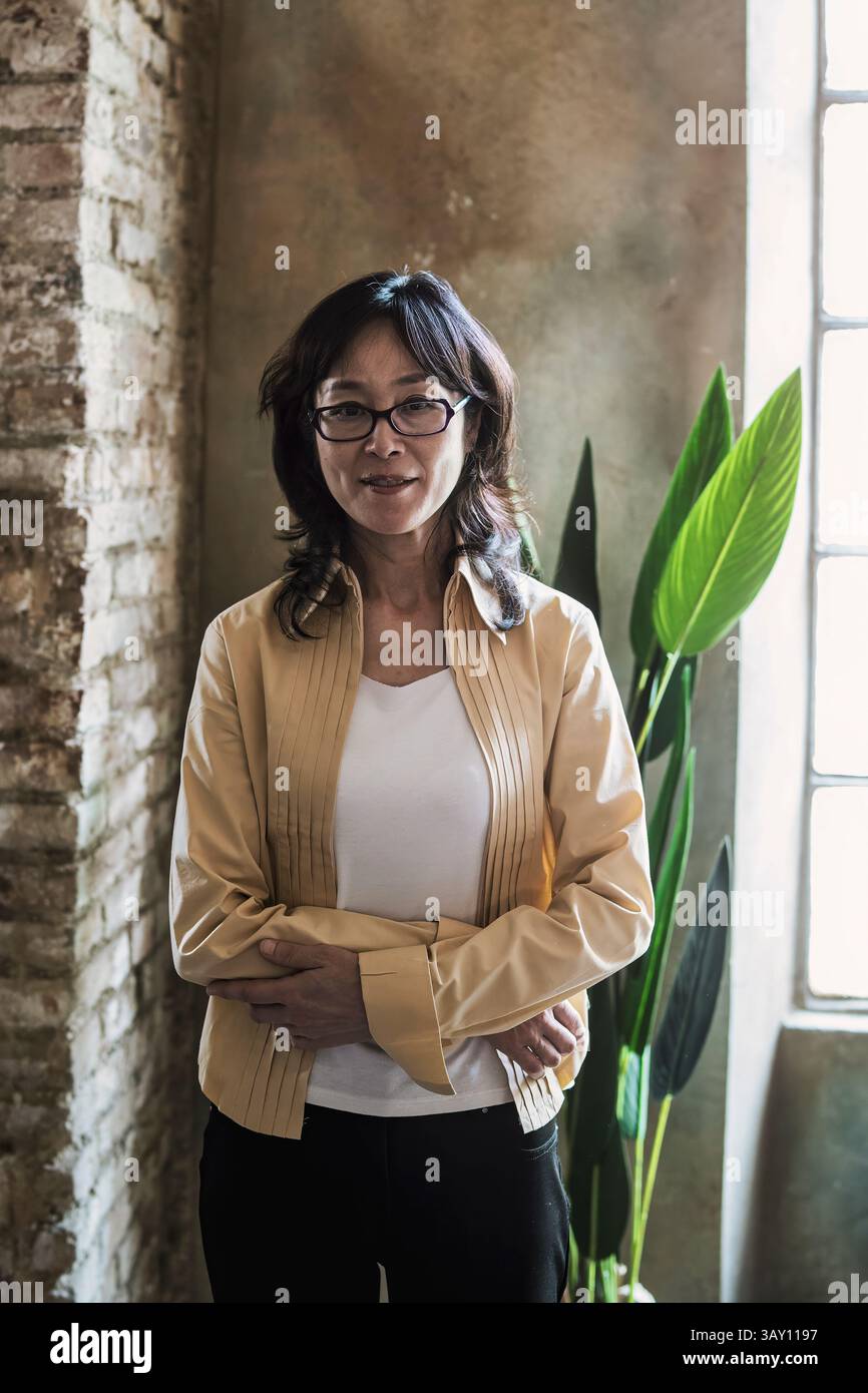 Ritratto di donna asiatica matura in piedi vicino alla finestra in un elegante spazio di lavoro con luce del giorno morbida ed espressione sicura Foto Stock