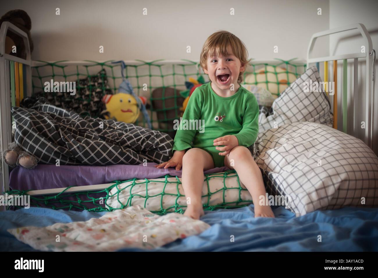 Un bambino che indossa una camicia verde si siede su un letto con vari cuscini e giocattoli, ridendo felicemente. La camera è luminosa e invitante, a creare un divertimento Foto Stock
