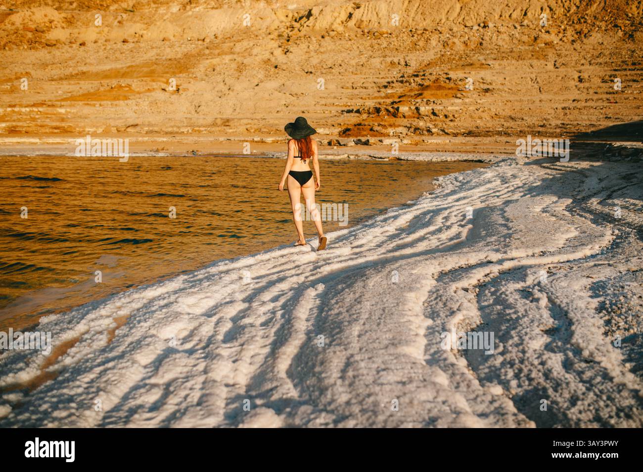 Vista posteriore di una donna irriconoscibile in bikini e cappello a teglia larga che cammina lungo le formazioni saline del Mar morto in Giordania. Foto Stock