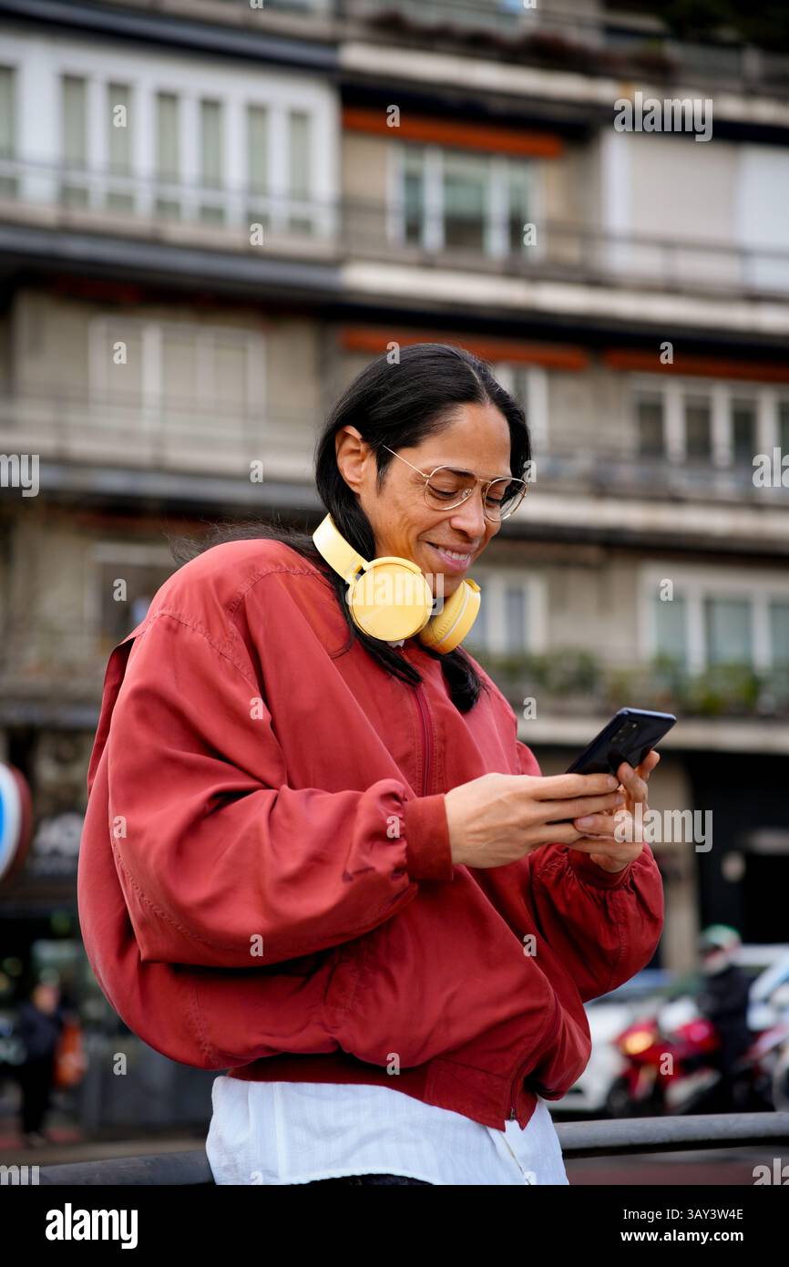 Un uomo con capelli lunghi e grandi occhiali con una giacca rossa e cuffie gialle è fuori a guardare uno smartphone, con un edificio urbano nella ba Foto Stock