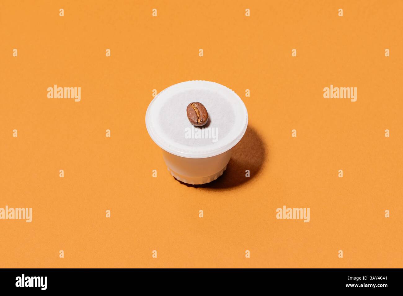 Una capsula di caffè biodegradabile posizionata su un vivace sfondo arancione, sormontata da un unico chicco di caffè, che mostra semplicità e modernità Foto Stock