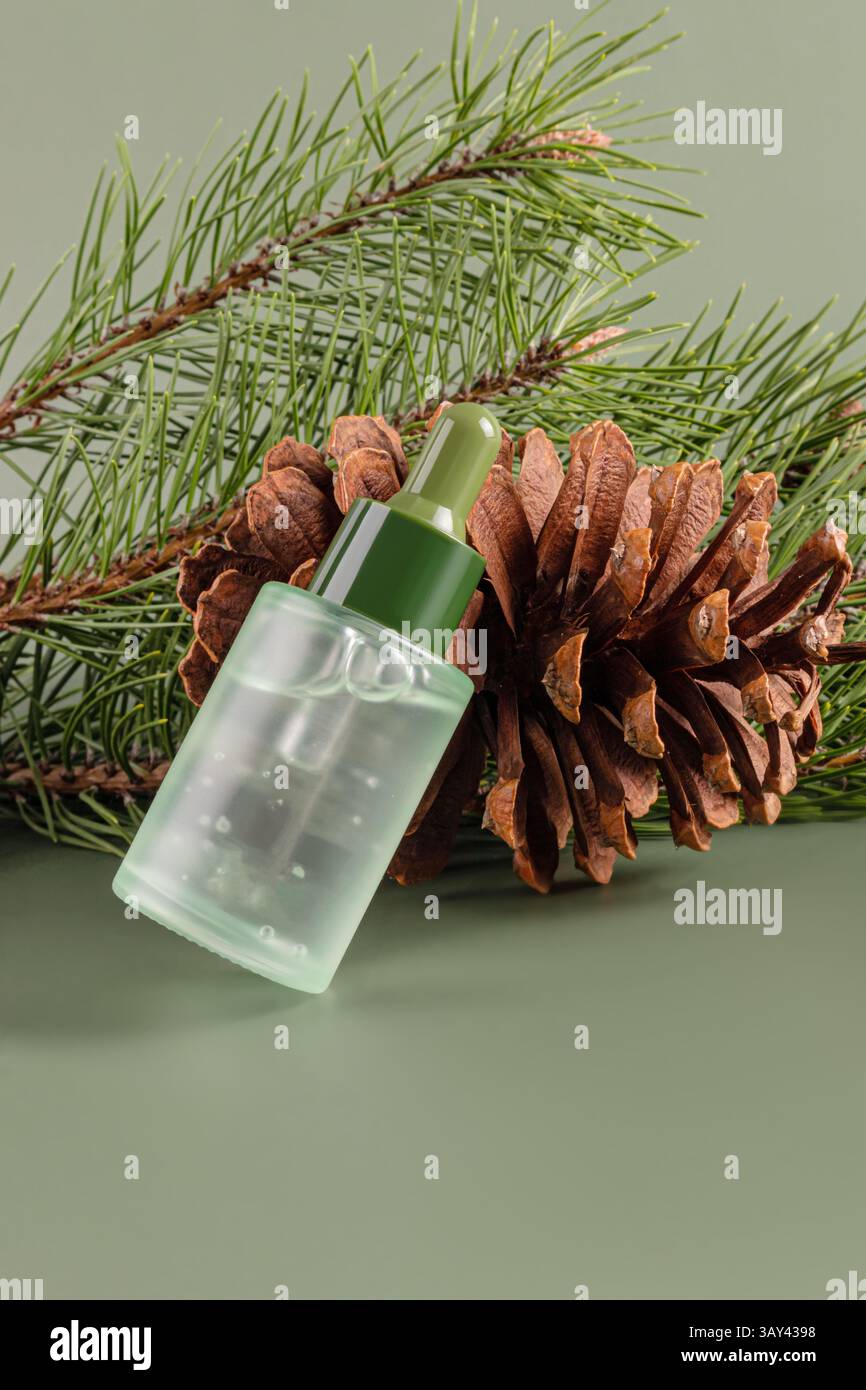 Vista ravvicinata di una bottiglia trasparente di olio essenziale di conifere, abbinata a un cono di pino e aghi di pino su un morbido sfondo verde. Foto Stock