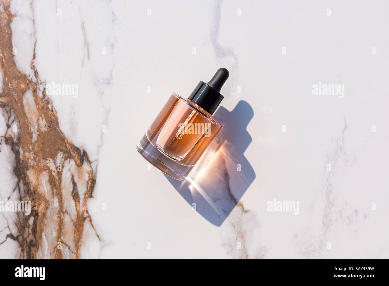 Un elegante flacone contagocce in vetro con liquido ambrato che getta un'ombra su una superficie in marmo. La luce del sole ne esalta l'aspetto elegante, ideale per la bellezza e il benessere Foto Stock