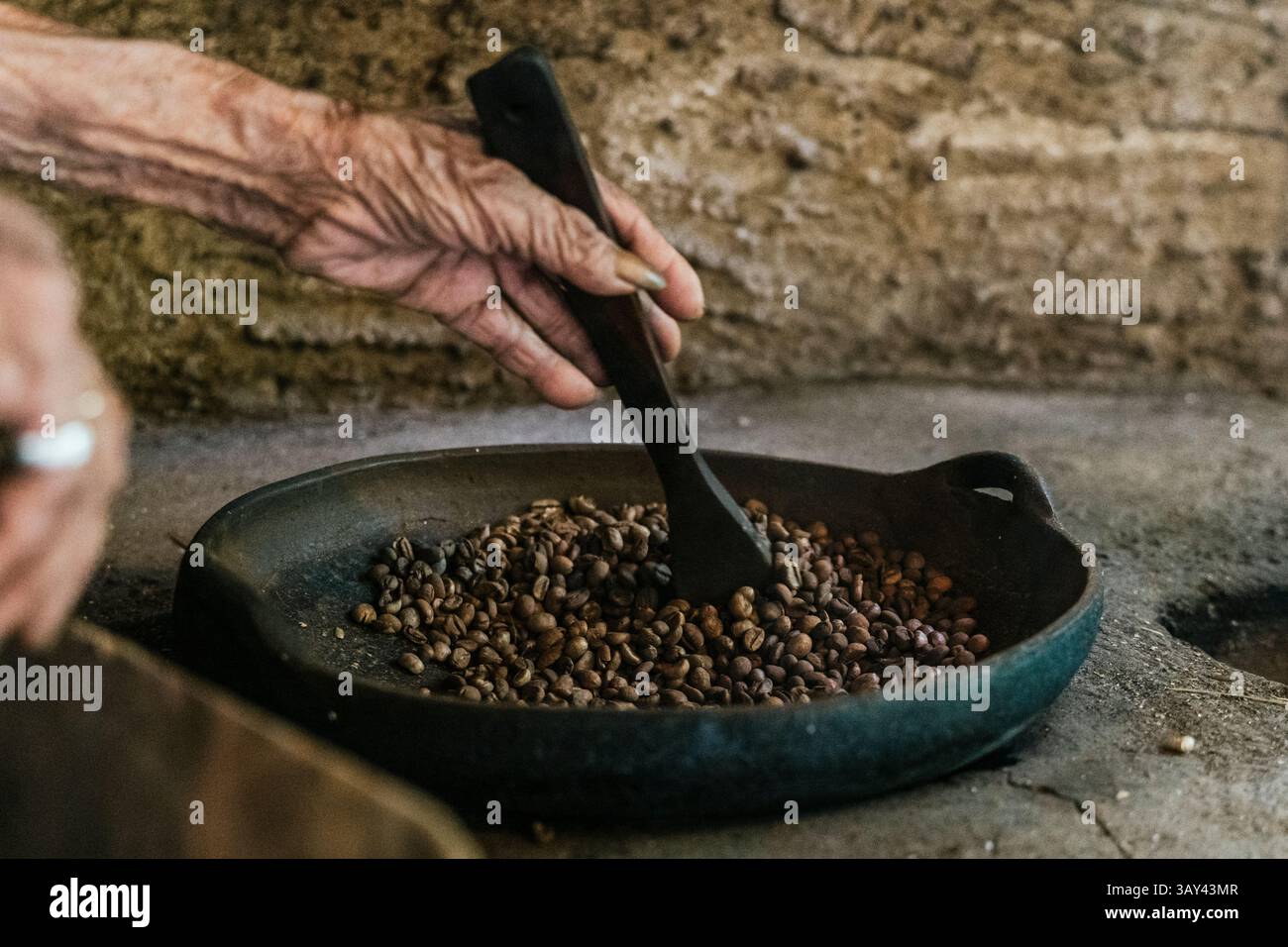 Una mano anziana mescola i chicchi di caffè balinesi in una padella, catturando l'essenza dei metodi di tostatura tradizionali in un ambiente rustico, evidenziando il culto Foto Stock