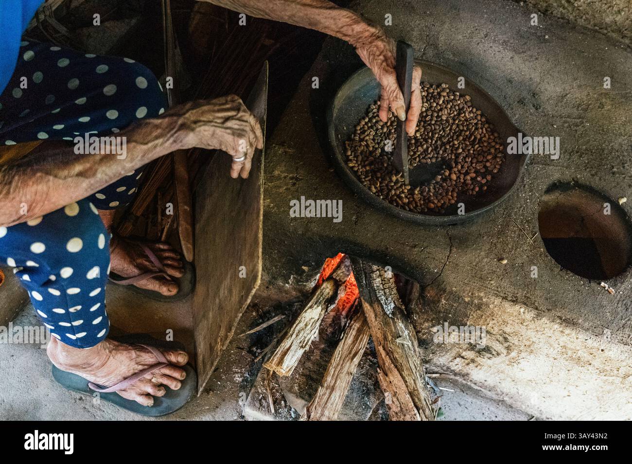 Primo piano di mani mescolando chicchi di caffè balinesi in una padella rustica su un fuoco a legna. La scena cattura i metodi tradizionali di tostatura del caffè, in alto Foto Stock