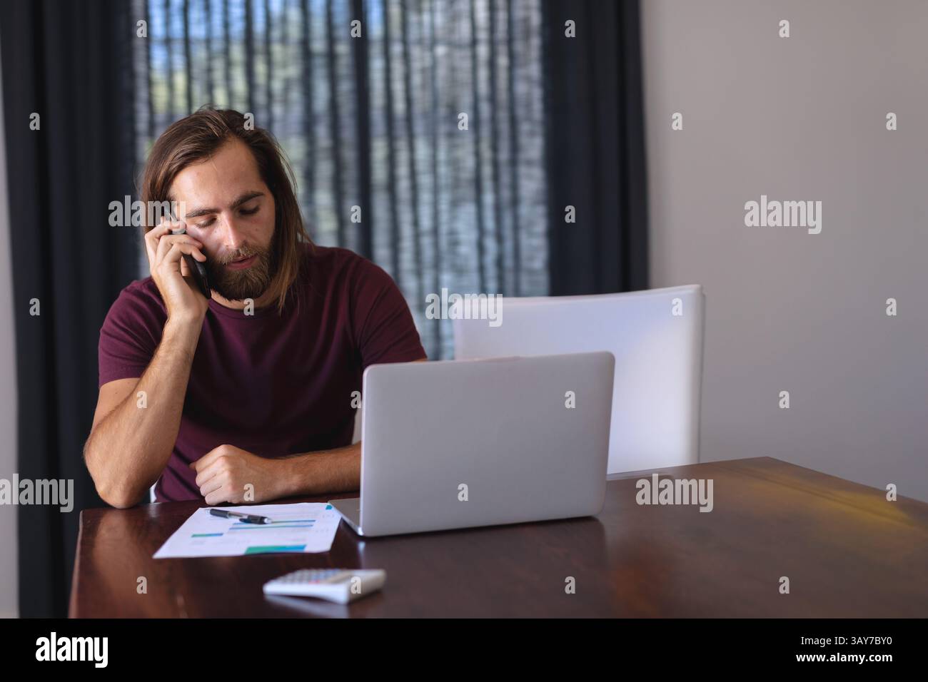 Giovane uomo adulto che parla su smartphone al tavolo dell'ufficio di casa, con laptop, grafici e calcolatrice Foto Stock