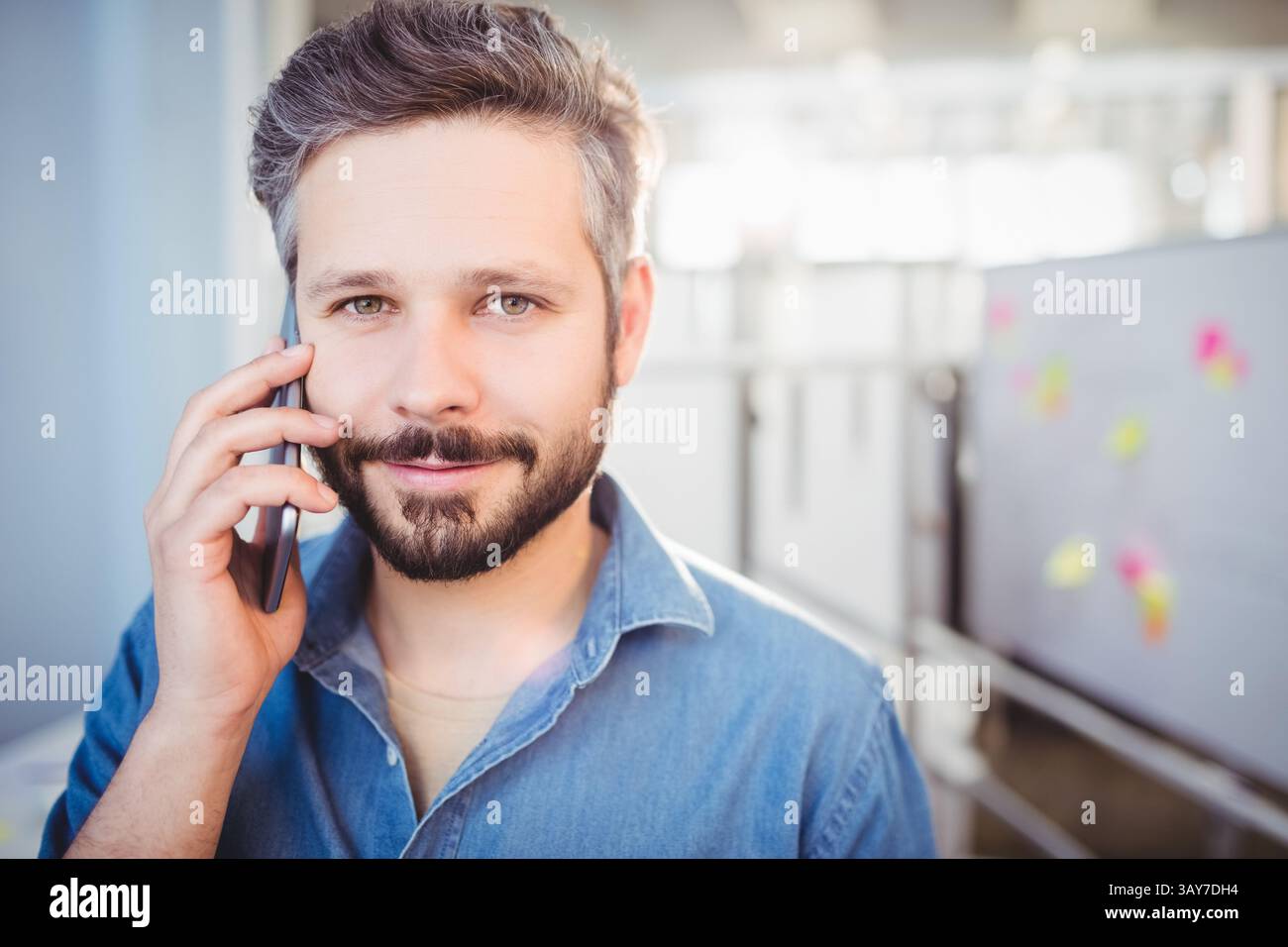 Uomo medio adulto in piedi in ufficio, tenendo lo smartphone all'orecchio e facendo contatto con gli occhi Foto Stock