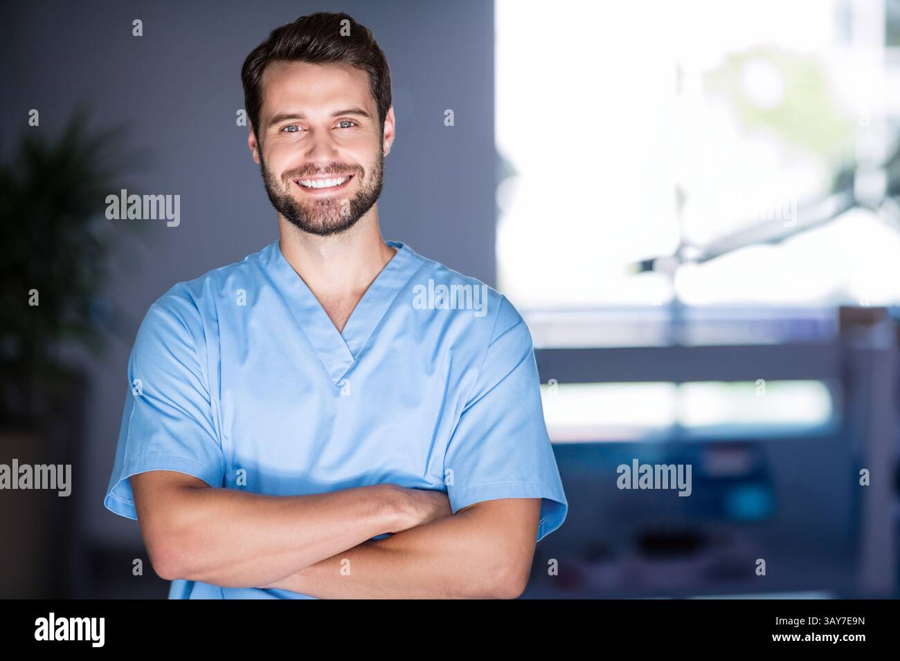 Uomo in piedi con le braccia incrociate in stanza d'ospedale, con scrub azzurri con piante e monitor Foto Stock