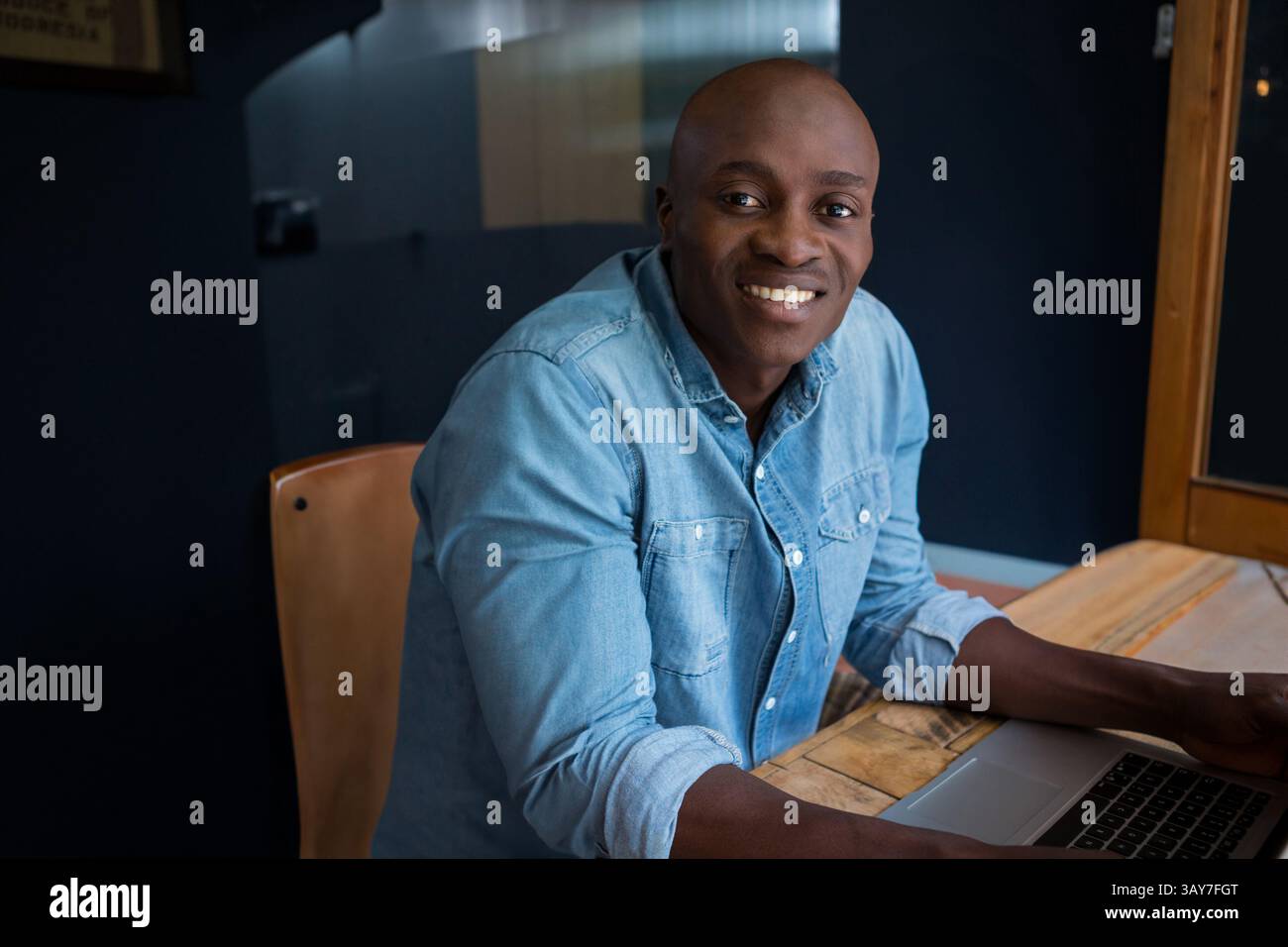 Giovane afroamericano adulto che utilizza un computer portatile in un moderno spazio di lavoro, con tavolo e sedia in legno Foto Stock