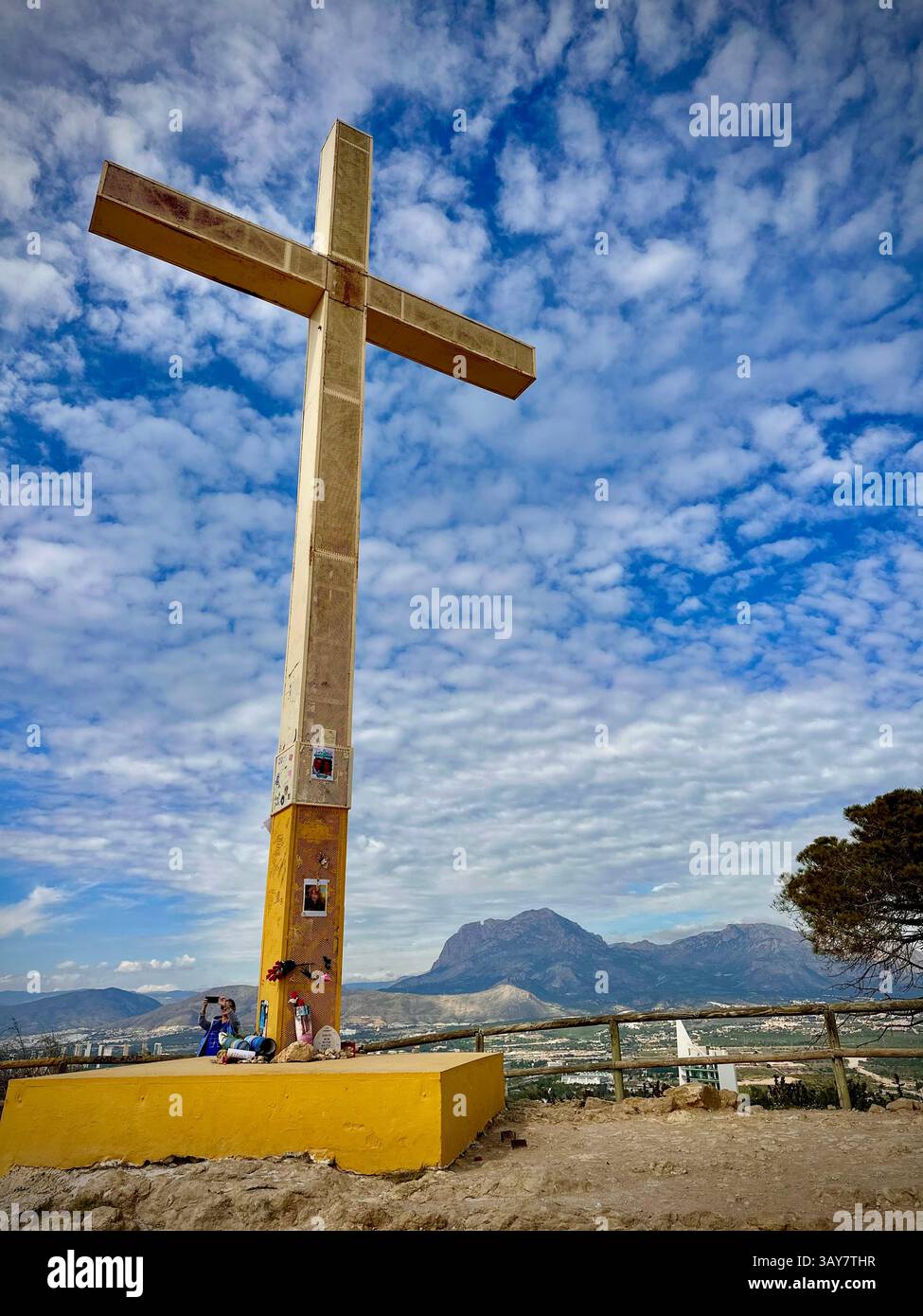 Una grande croce in cima alla collina si erge contro un cielo blu vibrante con nuvole sparse. Situato sopra un'ampia valle e circondato da cime montuose, questo tranquillo hotel - Immagine stock catturata con smartphone