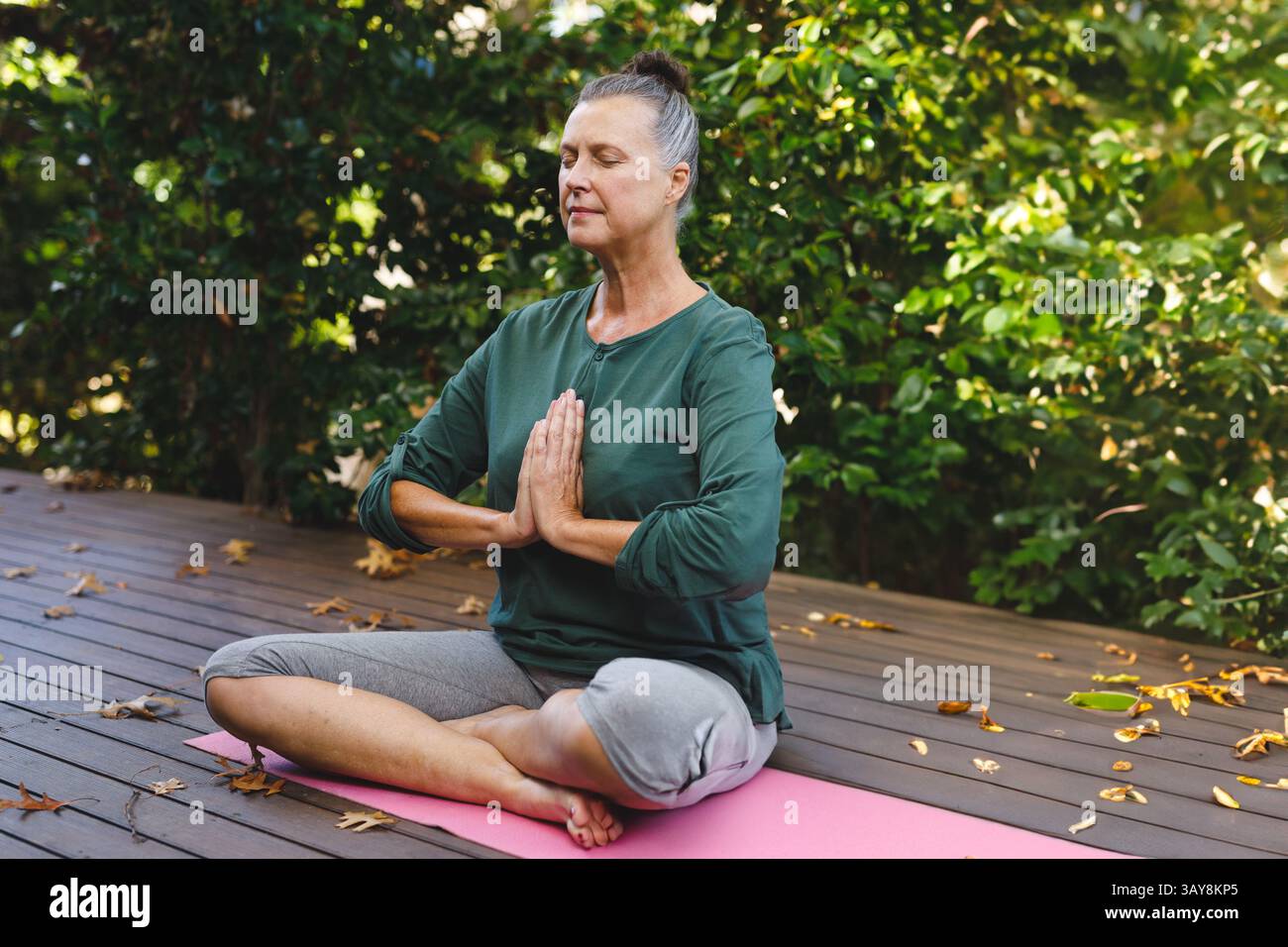 Donna anziana che pratica yoga sul ponte di legno nel giardino privato, con tappetino rosa e foglie sparse Foto Stock