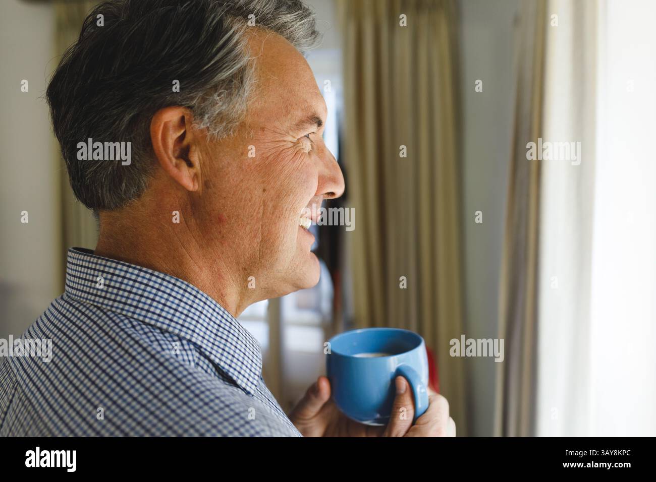 Uomo anziano che tiene la tazza di caffè in ceramica blu accanto alla finestra di casa, guarda fuori, copia spazio Foto Stock