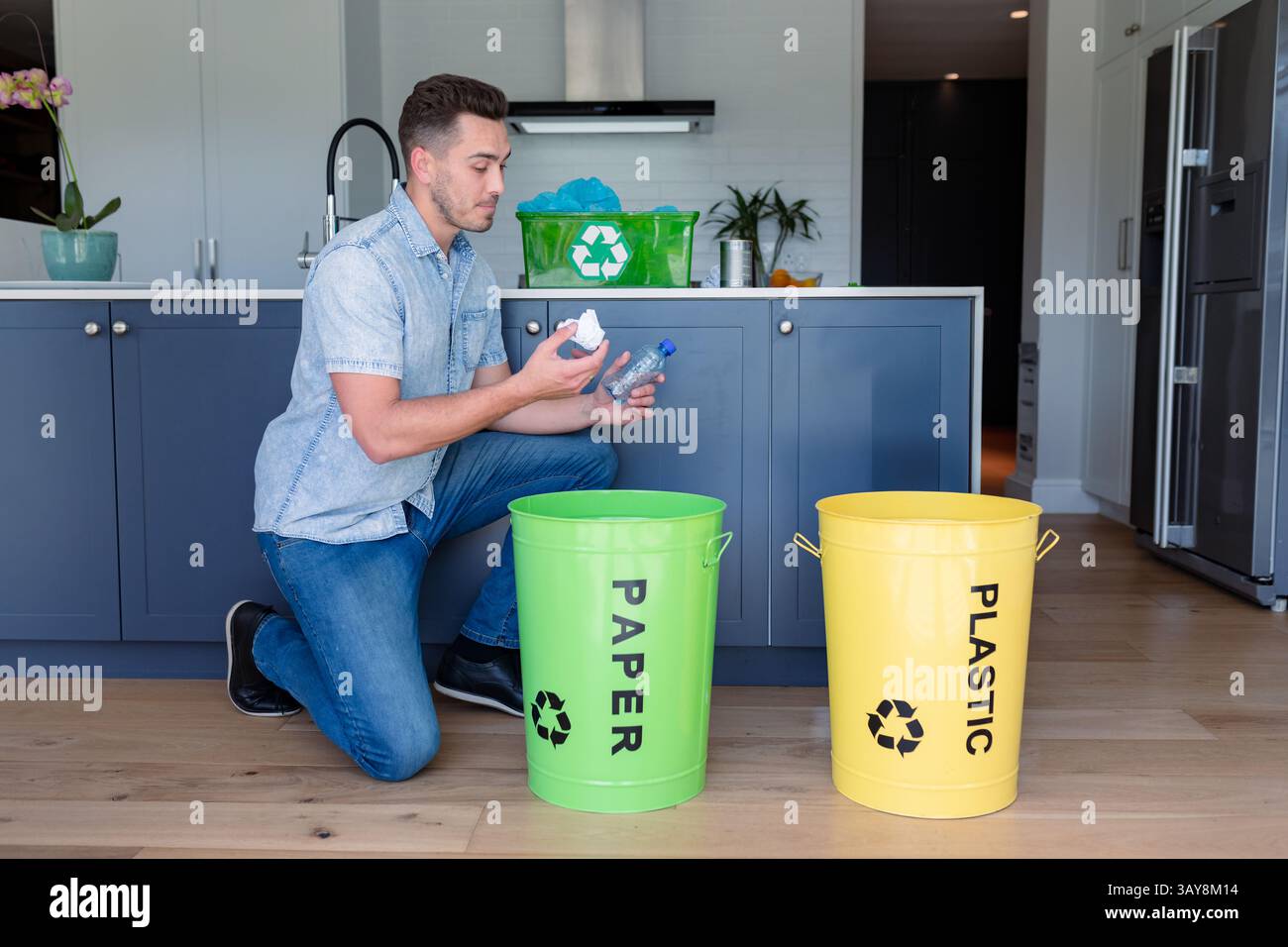 Giovane adulto che si inginocchia nella cucina di casa, smistando bottiglia e carta in contenitori di carta e plastica Foto Stock