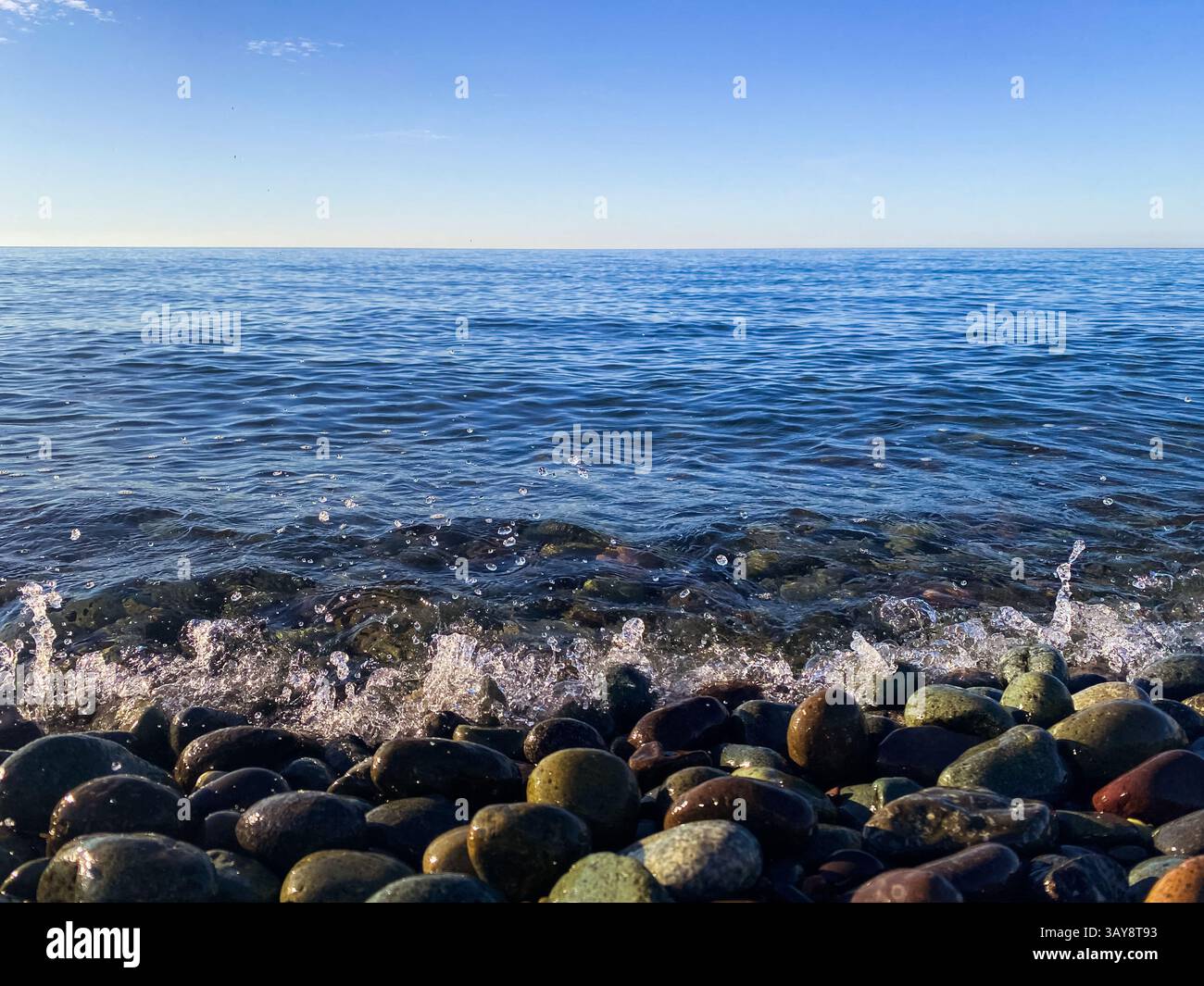Primo piano di onde oceaniche che spruzzano su ciottoli lisci sotto un cielo blu brillante, creando una tranquilla scena costiera. - Immagine stock catturata con smartphone