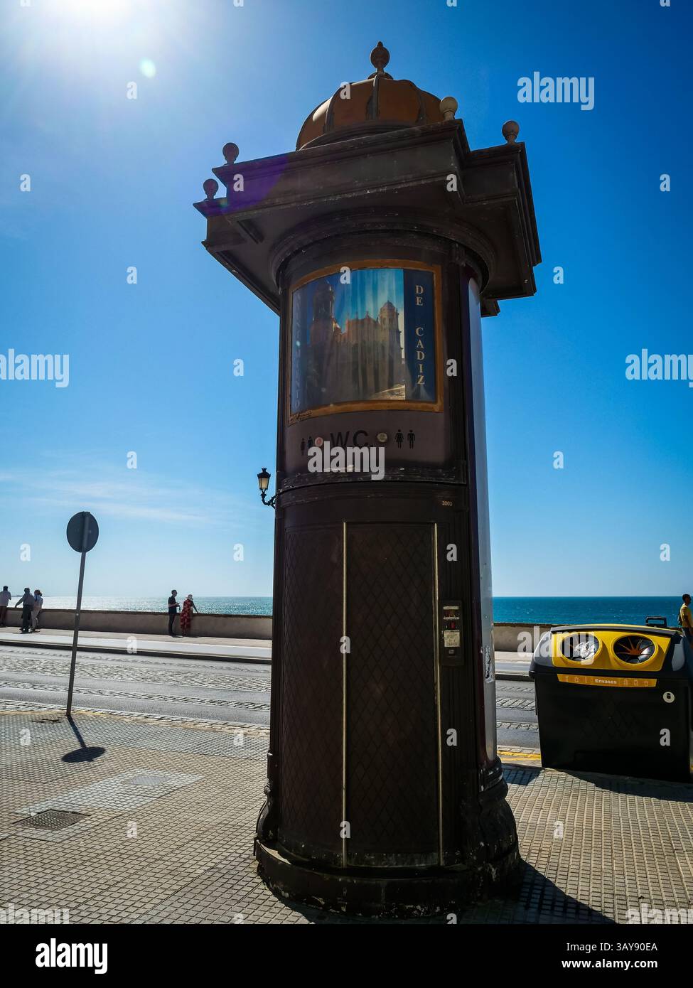 Un chiosco pubblico con servizi igienici distintivi e decorati si erge in modo prominente su un'area lastricata a Cadice, in Spagna, con il cielo blu brillante e l'oceano nel dorso Foto Stock