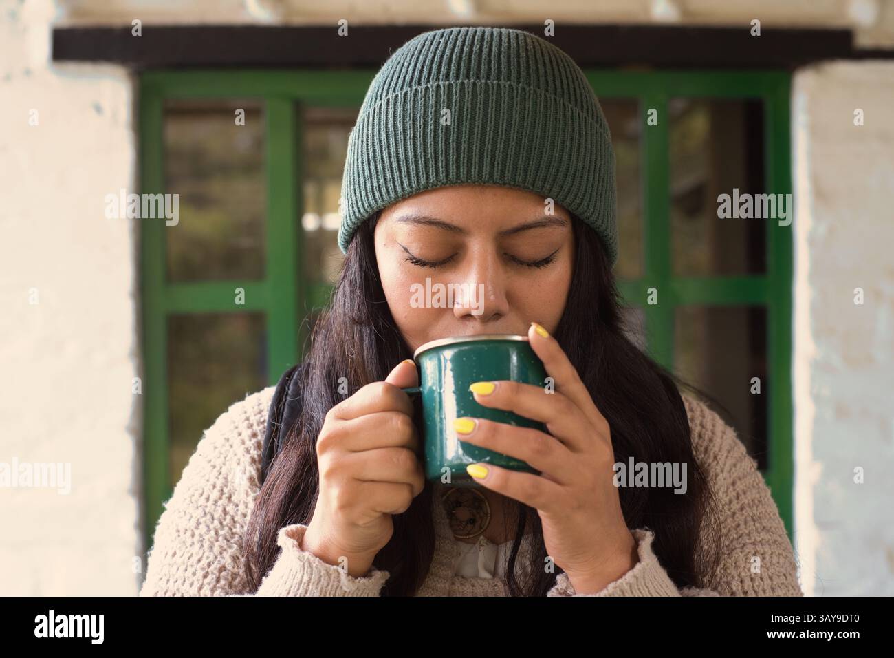 Una donna ama sorseggiare il suo delizioso drink, indossando un berretto accogliente, riflettendo sui momenti caldi trascorsi in casa Foto Stock