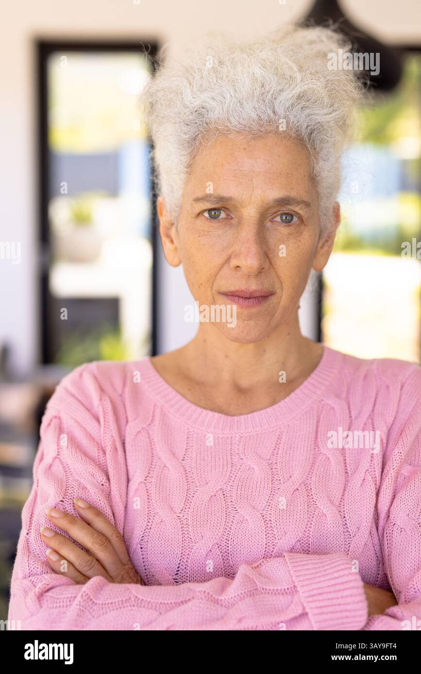 Donna anziana in piedi con le braccia incrociate in interni moderni, con un pullover rosa in maglia a cavo Foto Stock