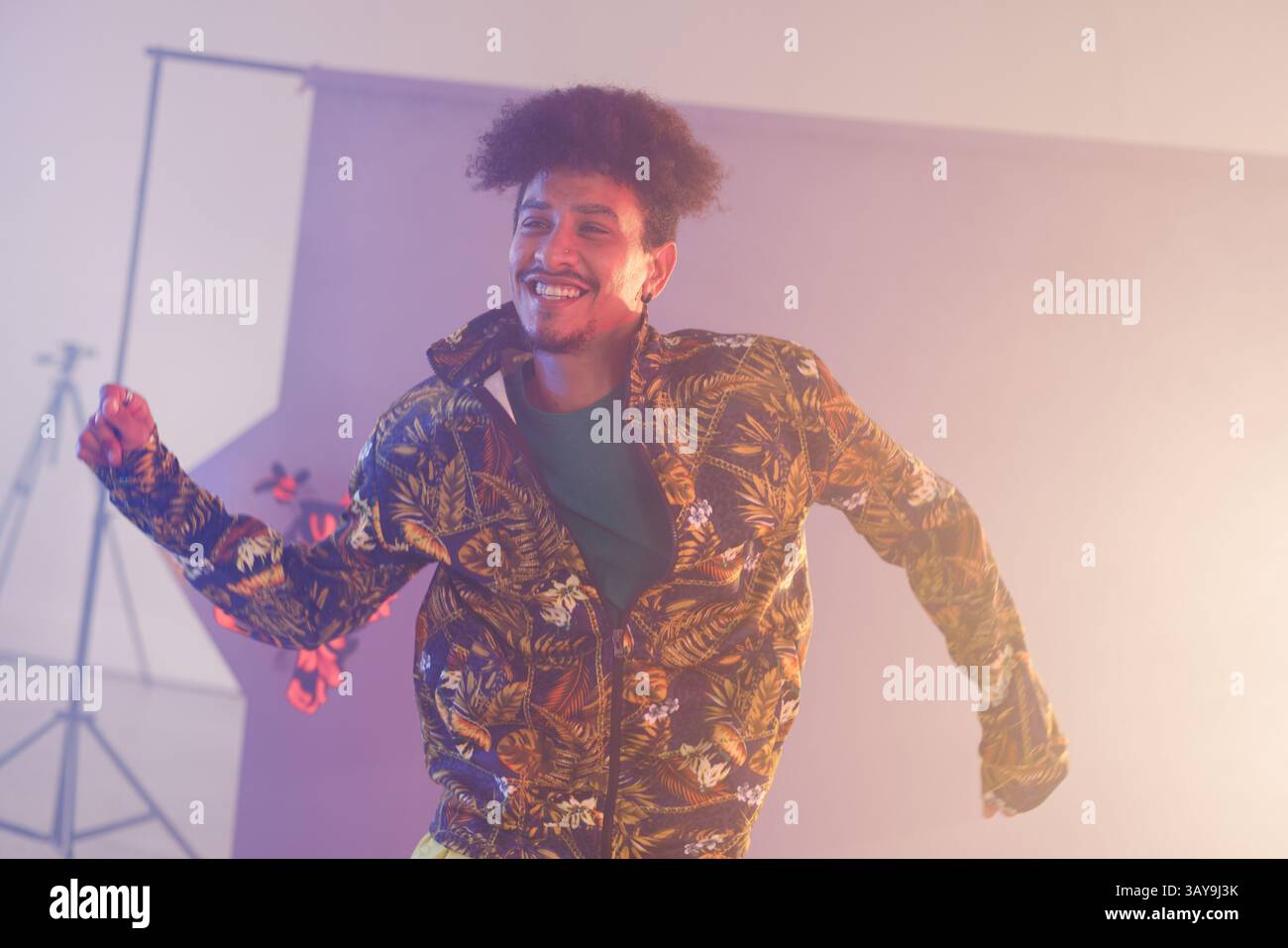 Uomo che balla e sorride nello studio fotografico, con giacca tropicale, treppiede e luci Foto Stock