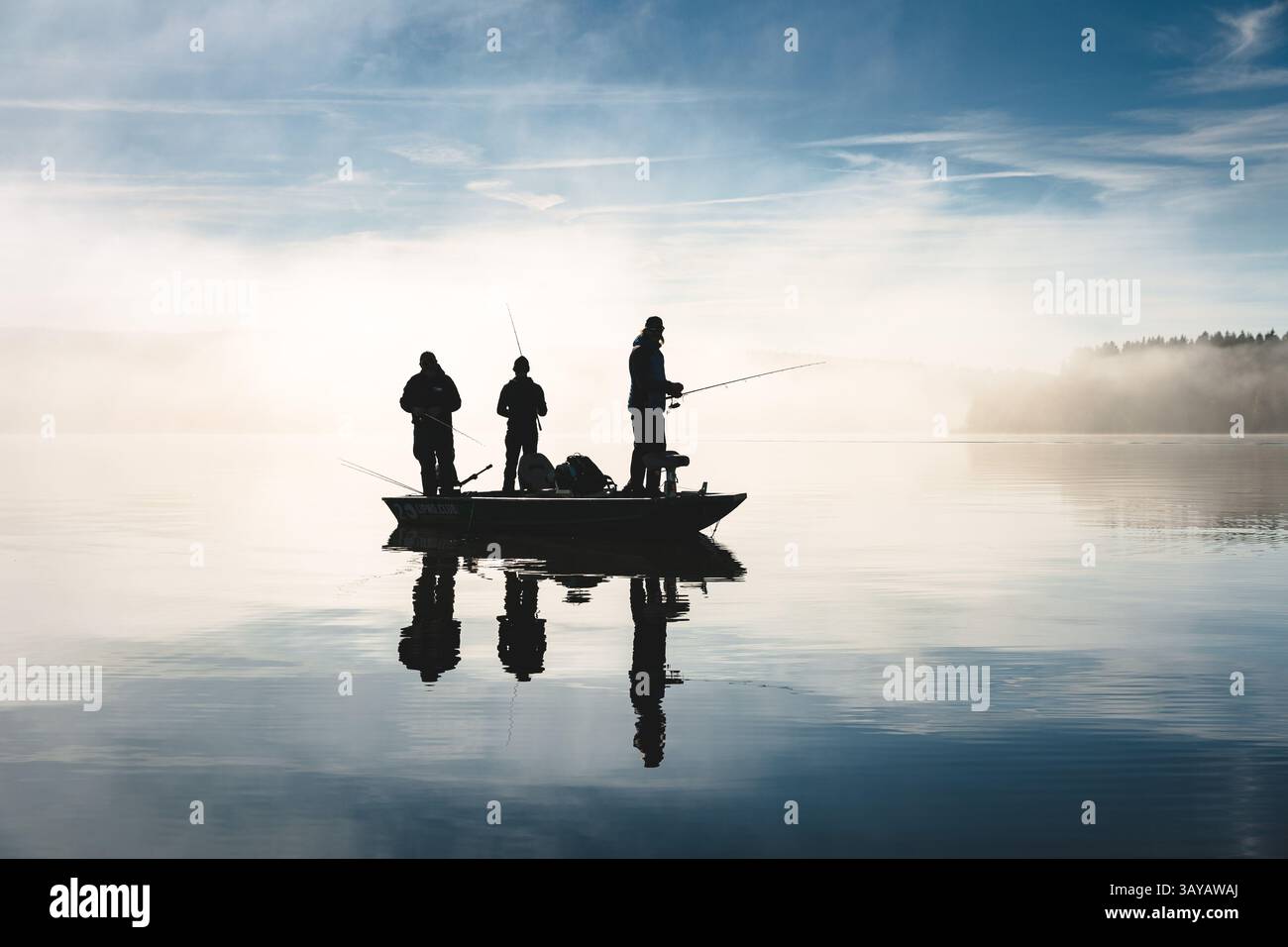 Pescatori in piedi su una barca su un lago in una giornata di sole con nebbia sullo sfondo, riflessi visibili sull'acqua, silhouette sulla retroilluminazione Foto Stock