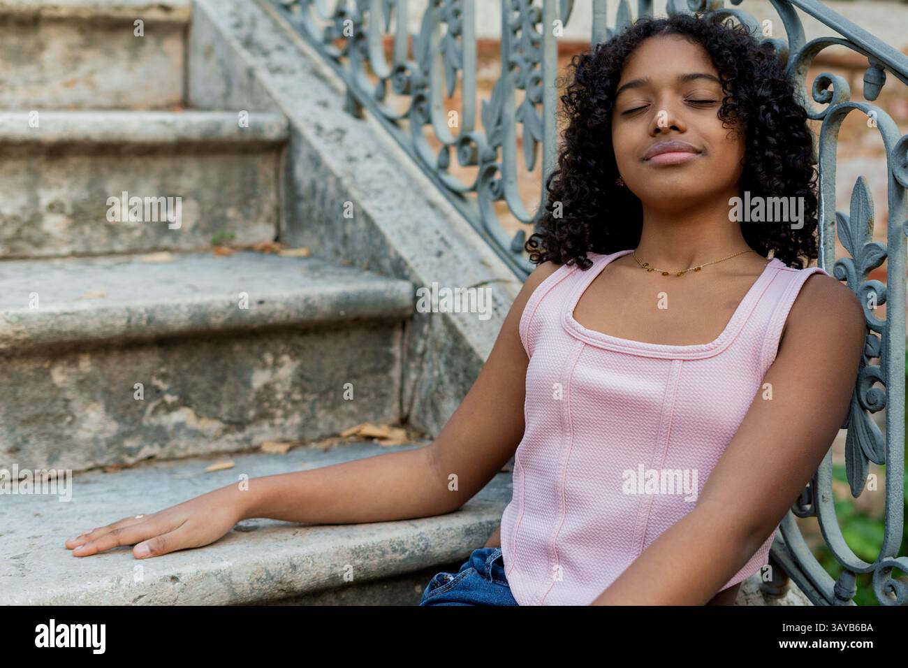 Giovane donna nera con capelli ricci e canotta rosa è seduta e rilassante a occhi chiusi su antiche scale di pietra accanto a una ringhiera in metallo Foto Stock Giovane donna nera con capelli ricci e canotta rosa è seduta e rilassante a occhi chiusi su antiche scale di pietra accanto a una ringhiera in metallo Foto Stock