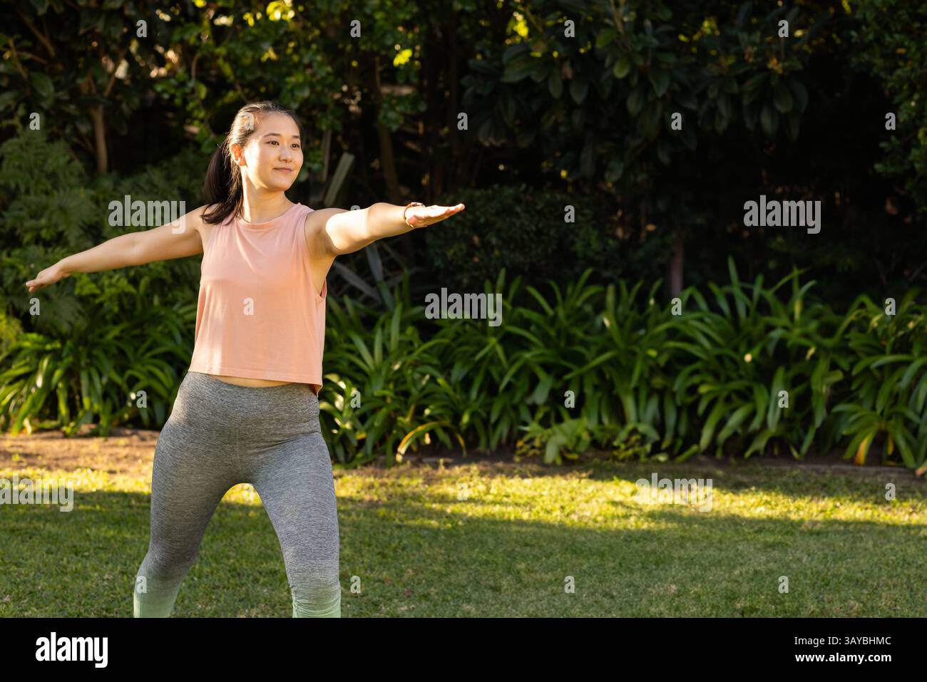 Donna asiatica di mezza età che pratica lo yoga Warrior II posa su un prato erboso, godendosi la calda luce del sole Foto Stock