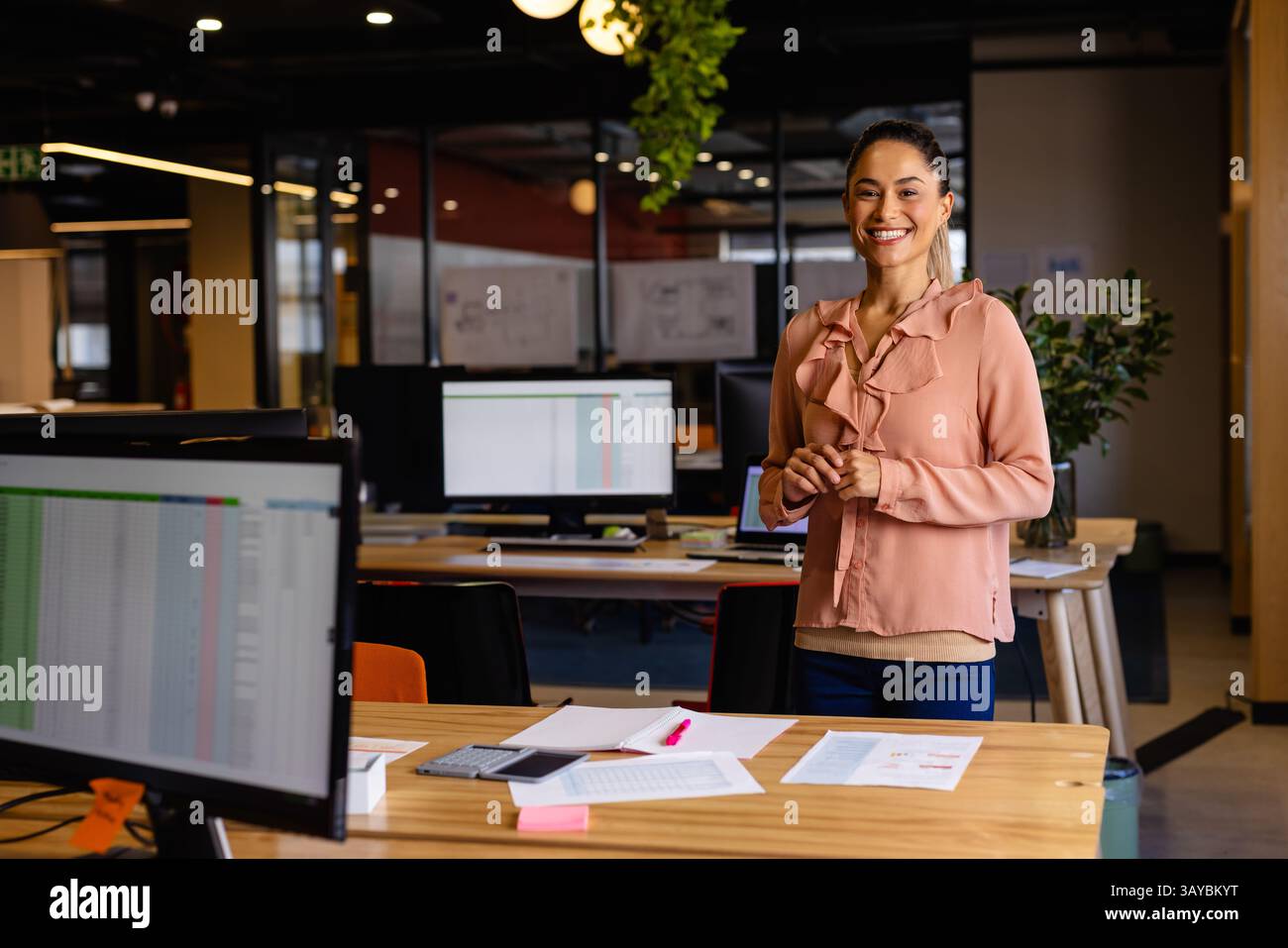 Donna in piedi e sorridente in un ufficio moderno, con monitor per computer e smartphone, spazio di copia Foto Stock
