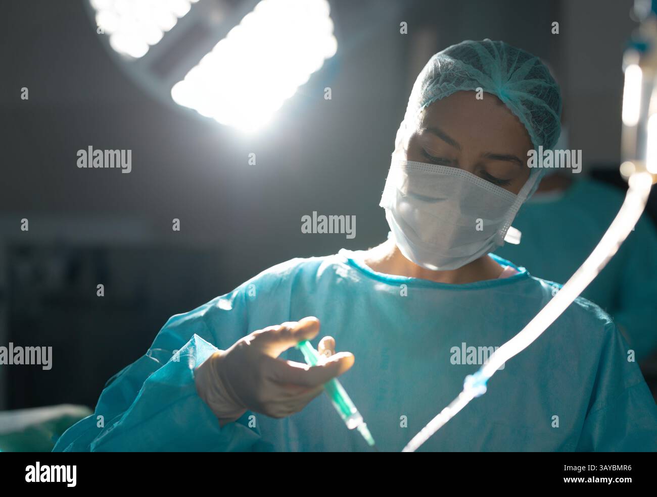 Giovane chirurgo femminile con maschera per preparare la siringa sotto la lampada chirurgica con tubi in sala operatoria Foto Stock