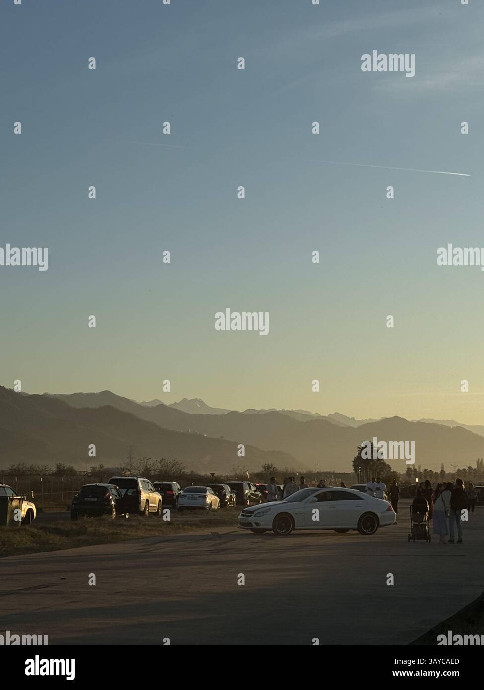Questa immagine mostra una scena a bordo strada durante il tramonto. Diverse auto parcheggiate e persone, tra cui un gruppo con un passeggino, sono visibili in primo piano Foto Stock Questa immagine mostra una scena a bordo strada durante il tramonto. Diverse auto parcheggiate e persone, tra cui un gruppo con un passeggino, sono visibili in primo piano Foto Stock