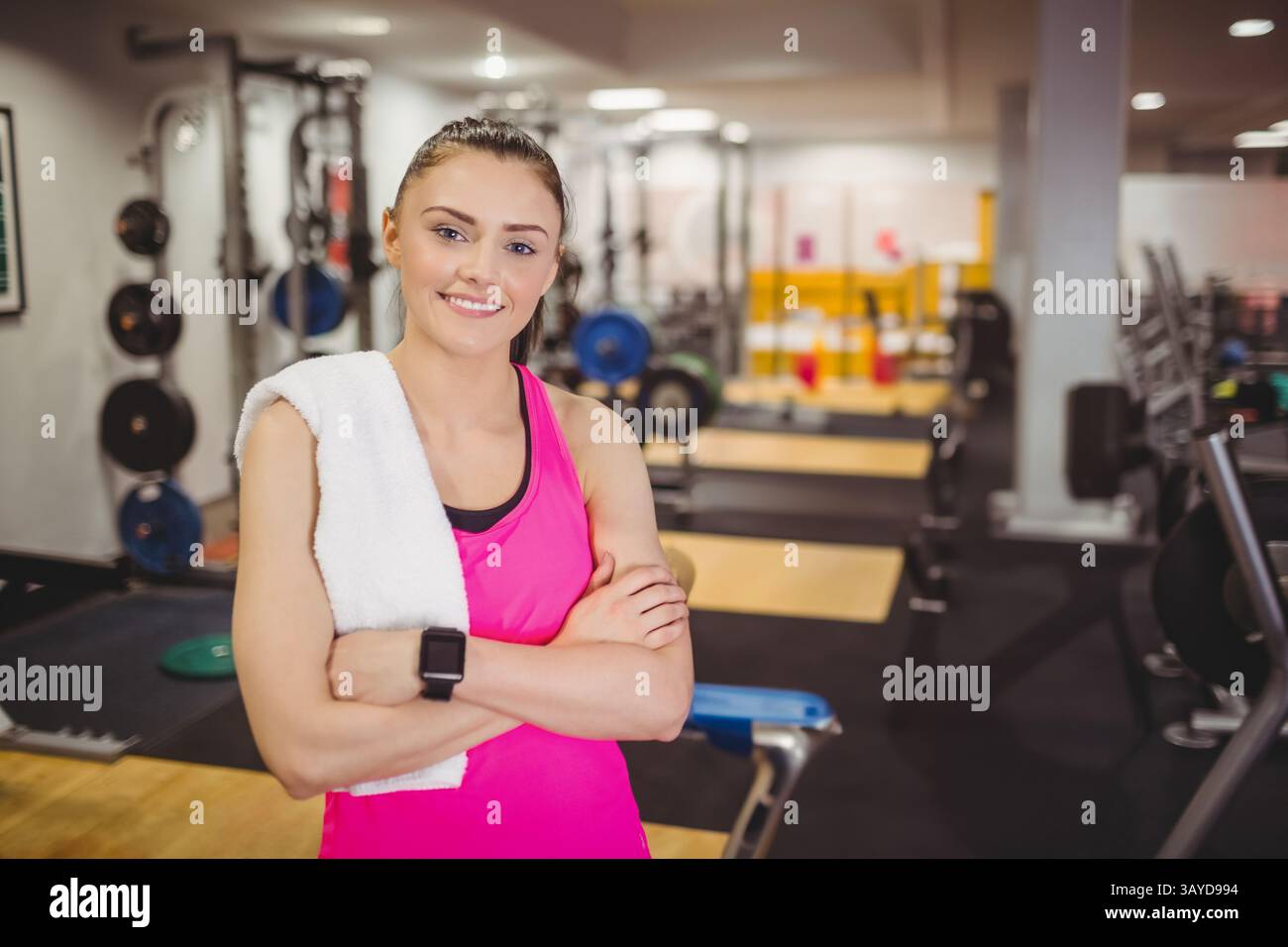 Giovane donna adulta in piedi con le braccia incrociate in palestra, con un asciugamano e uno smartwatch Foto Stock Giovane donna adulta in piedi con le braccia incrociate in palestra, con un asciugamano e uno smartwatch Foto Stock