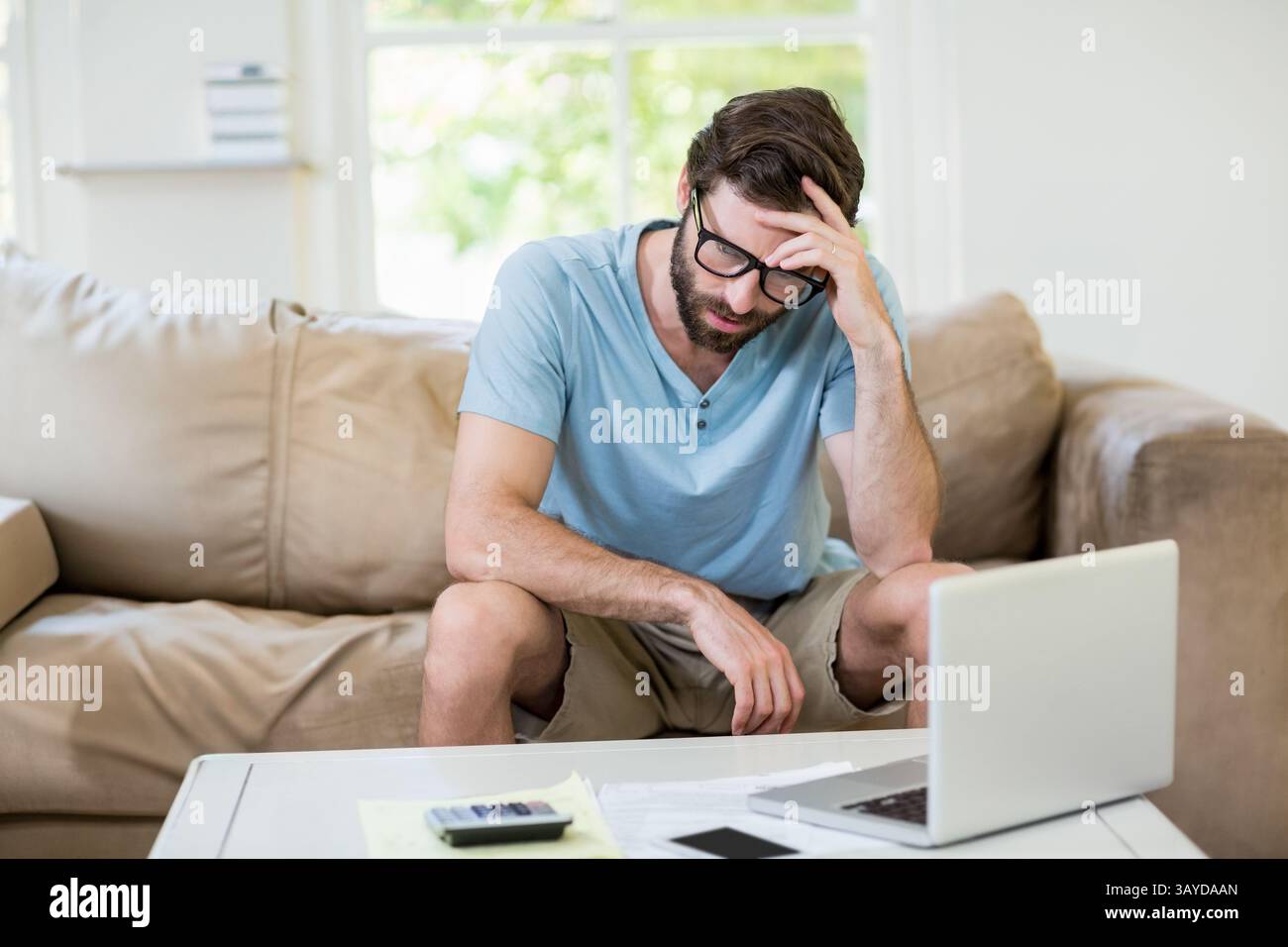 Uomo seduto sul divano in soggiorno, che si concentra sul computer portatile con la calcolatrice di documenti e smartphone Foto Stock