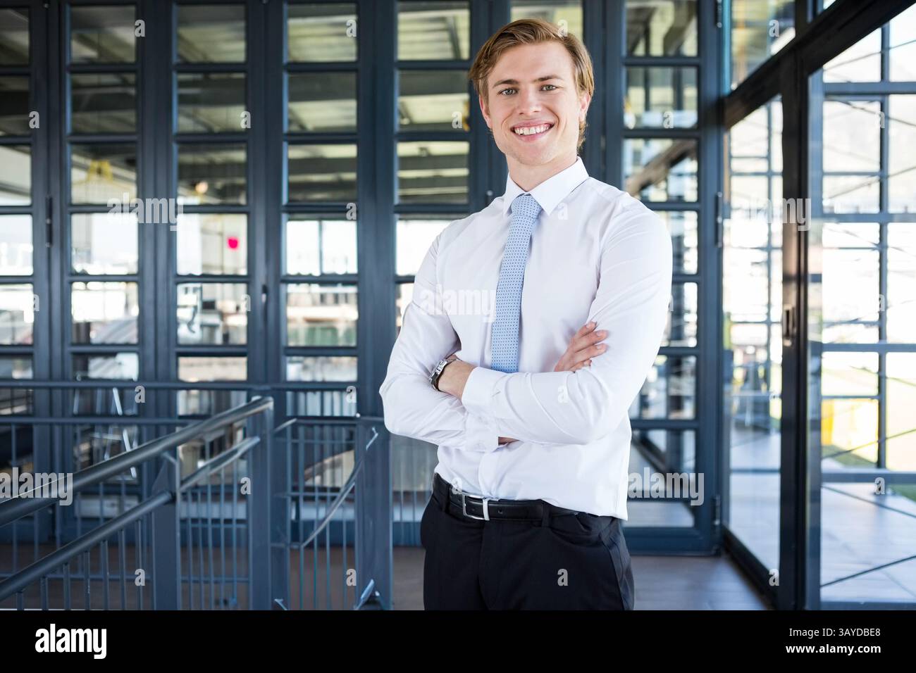 Uomo in piedi nel corridoio mezzanino dell'ufficio con le braccia incrociate e sorridenti, che mostra travi metalliche Foto Stock