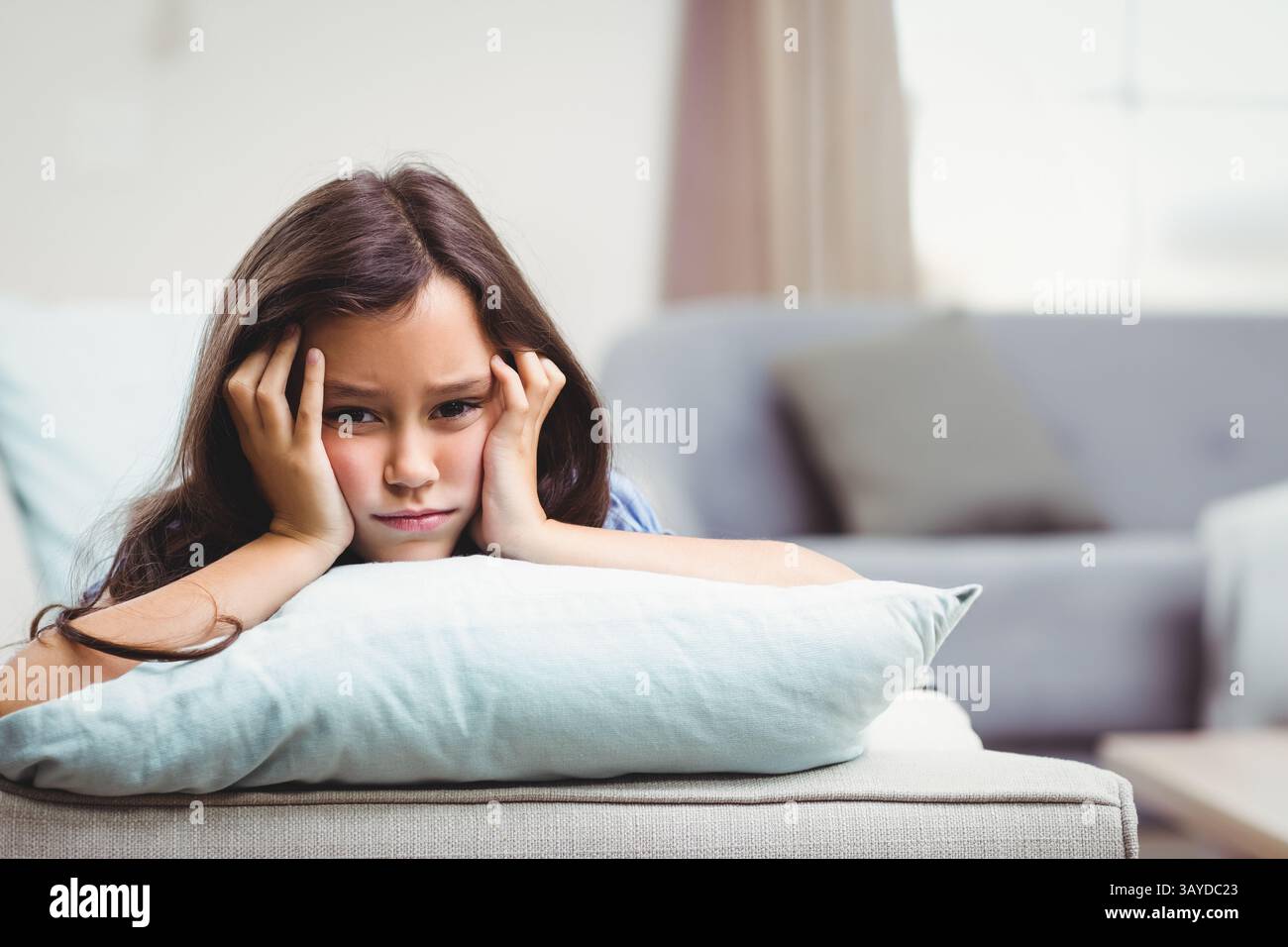 Un bambino che si appoggia su un cuscino blu sul divano neutro nel soggiorno e ha un aspetto premuroso Foto Stock