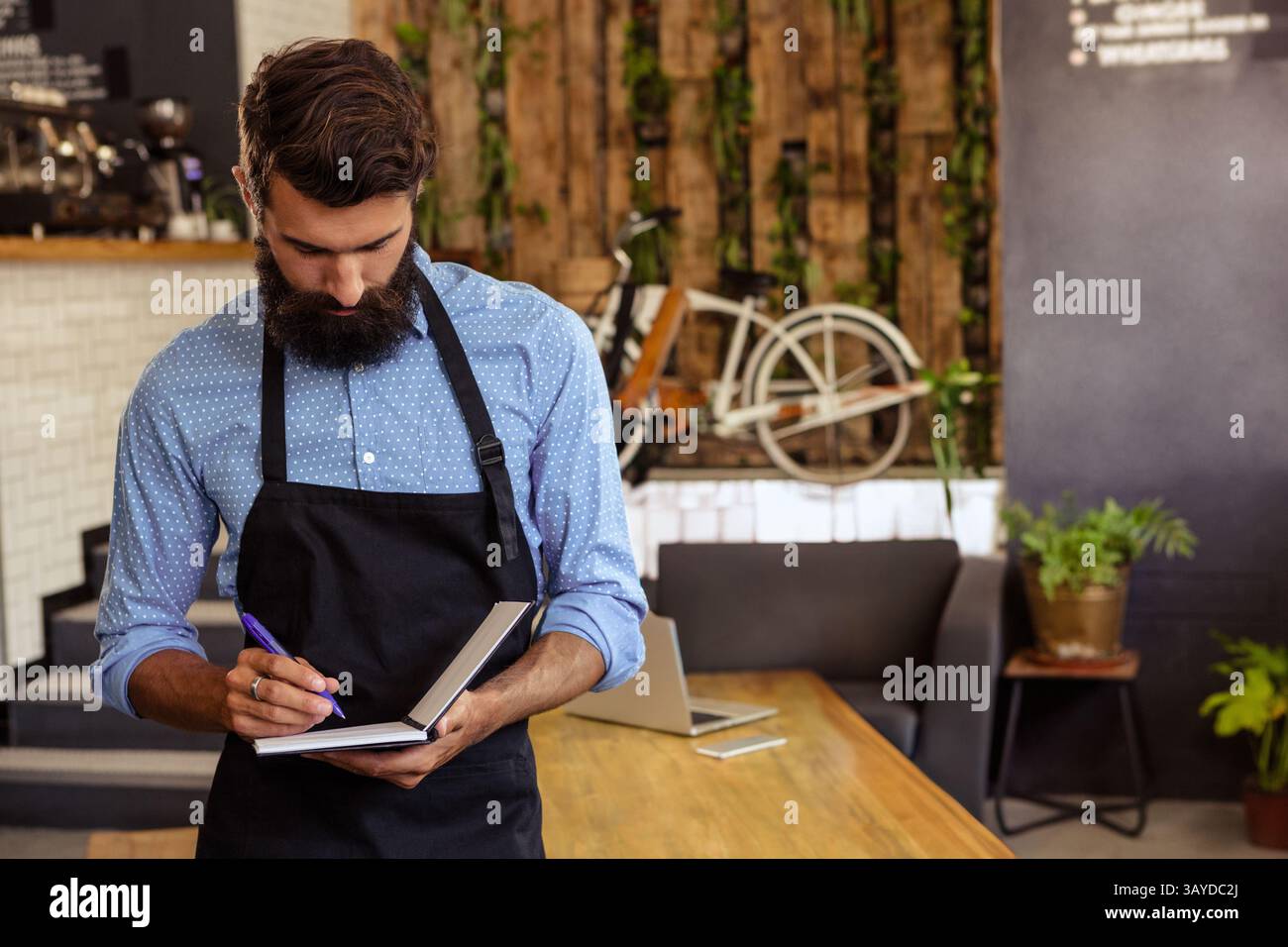 Giovane barista maschio che scrive appunti al tavolo di legno all'interno del rustico café, con taccuino e penna Foto Stock