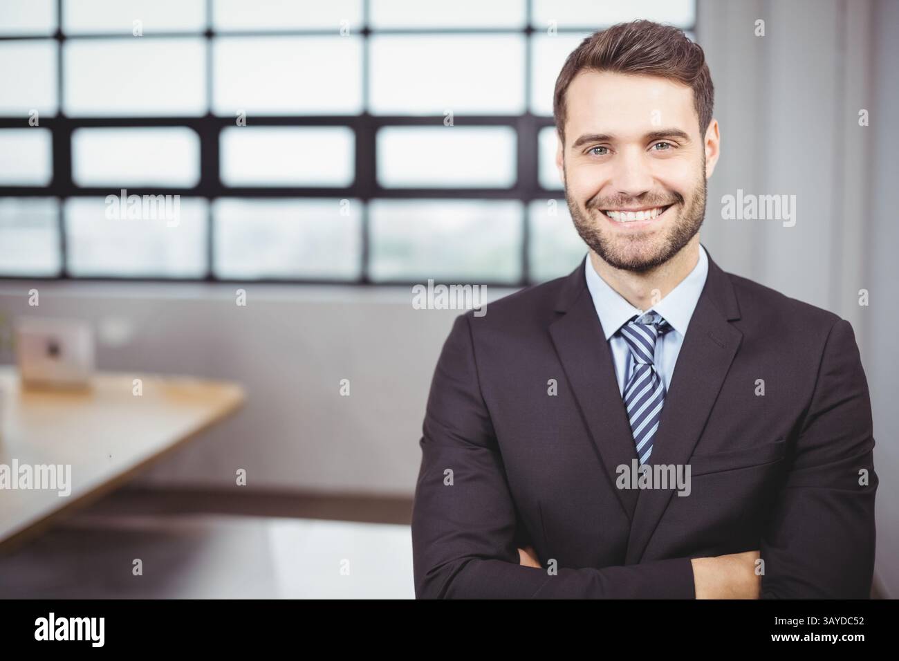 Uomo d'affari in piedi alla scrivania che incrocia le braccia in ufficio, con cravatta nera e finestra Foto Stock