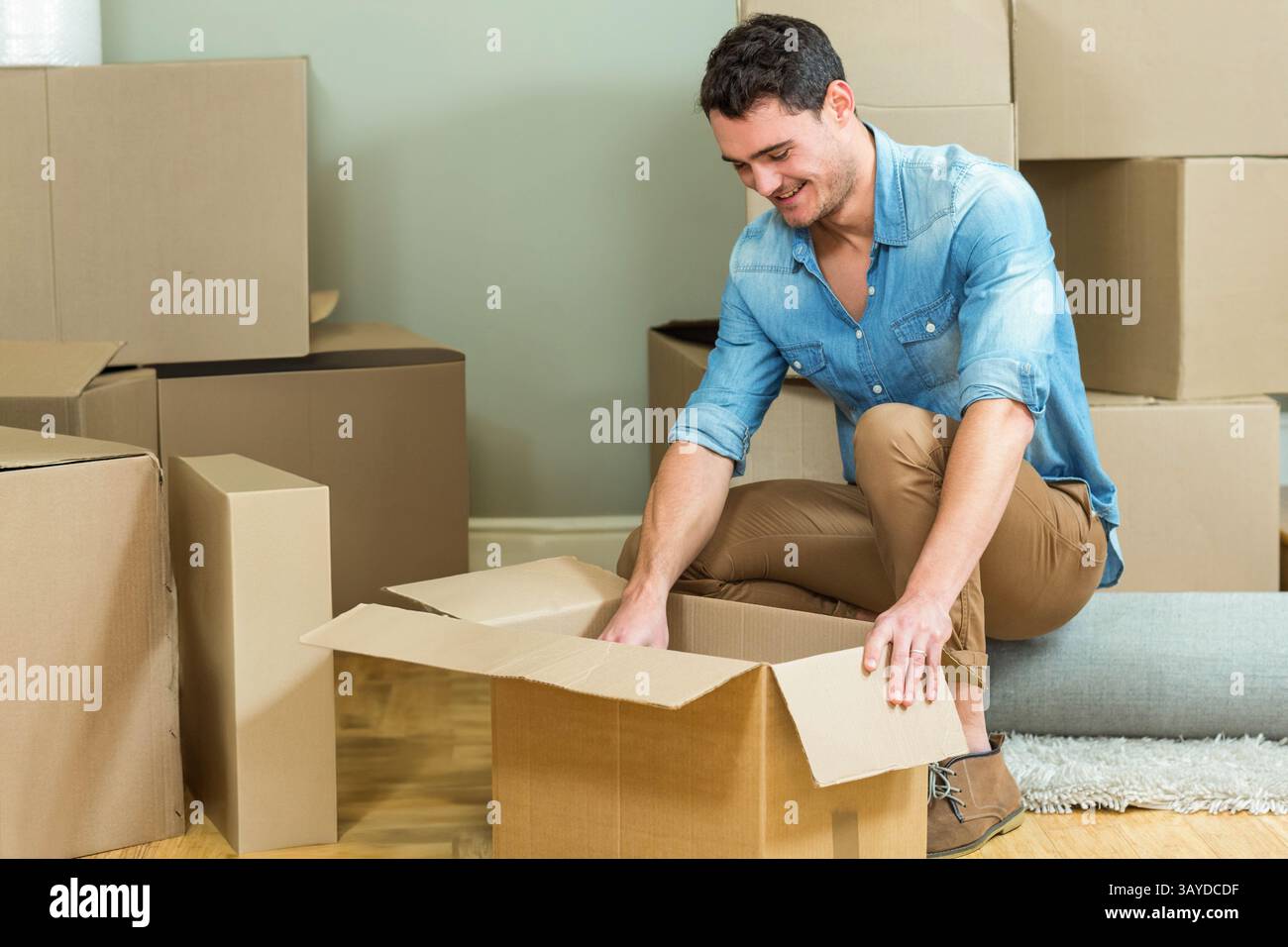 Uomo inginocchiato disimballando una scatola di cartone su pavimento in legno nel soggiorno, con tappeto arrotolato, spazio per le copie Foto Stock