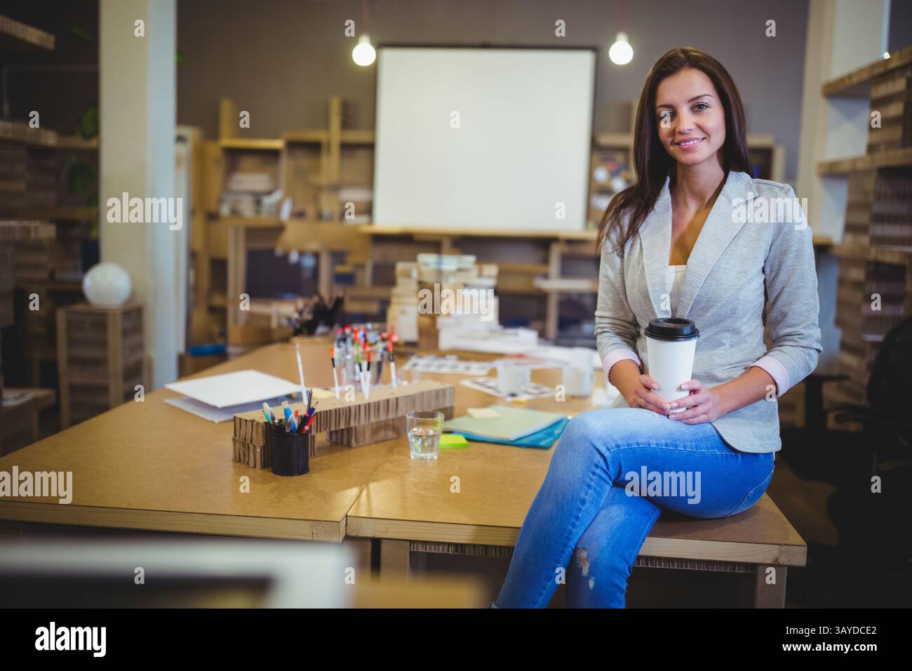 Donna seduta sul bordo del tavolo in uno studio di design con modelli in cartone, in cui si tiene una tazza di caffè, spazio fotocopie Foto Stock