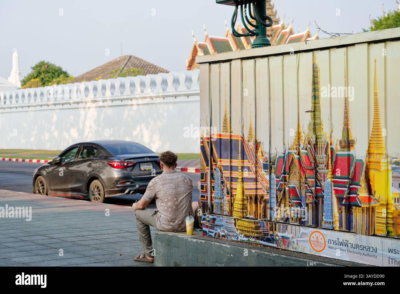 L'uomo si rilassa accanto a un colorato murale del Grand Palace di Bangkok, mentre i veri tetti dei templi sbirciano sopra le mura sullo sfondo Foto Stock