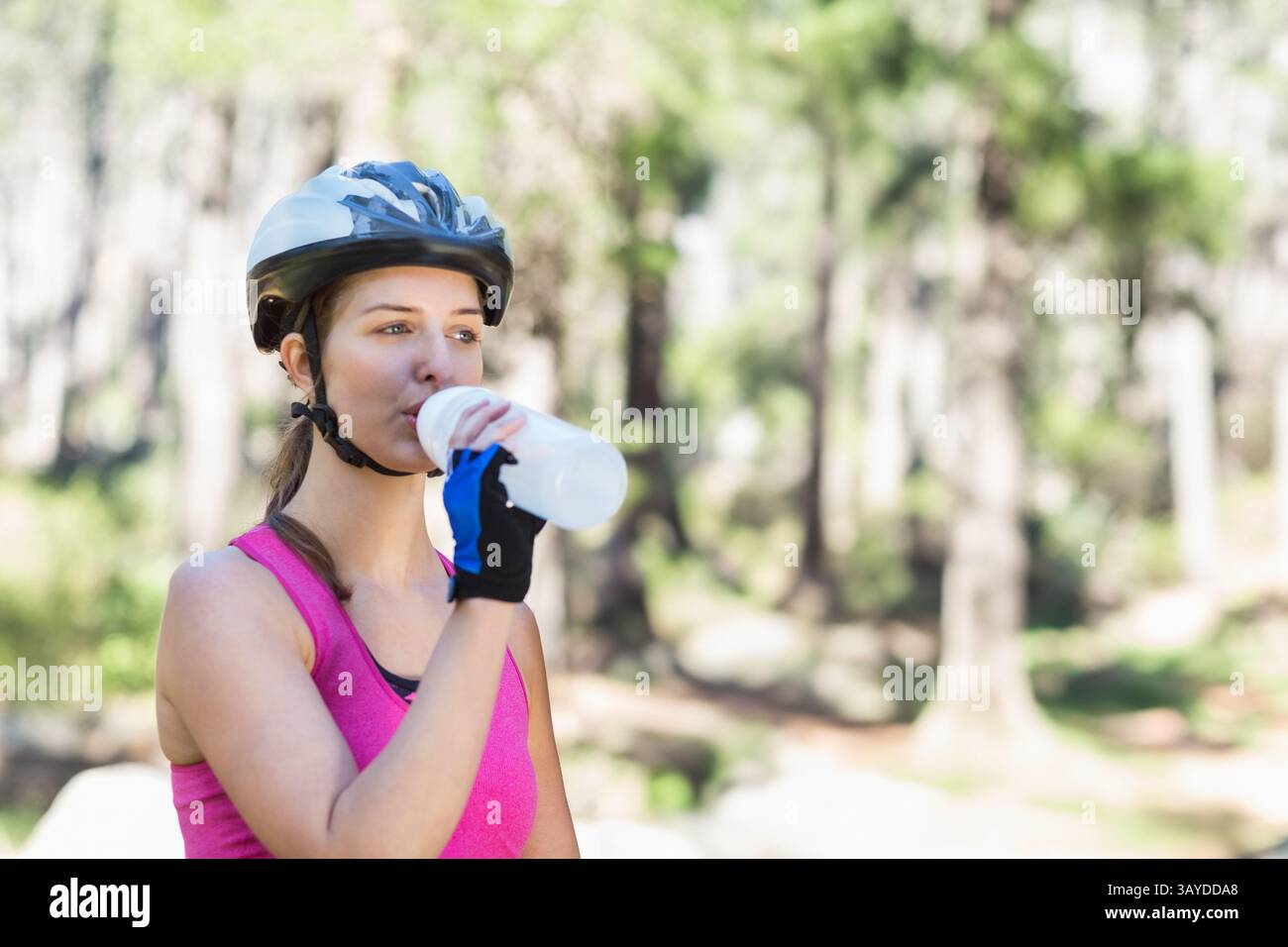 Ciclista donna che indossa il casco bevendo da una bottiglia d'acqua bianca su un sentiero nella foresta illuminato dal sole, spazio per copiare Foto Stock Ciclista donna che indossa il casco bevendo da una bottiglia d'acqua bianca su un sentiero nella foresta illuminato dal sole, spazio per copiare Foto Stock