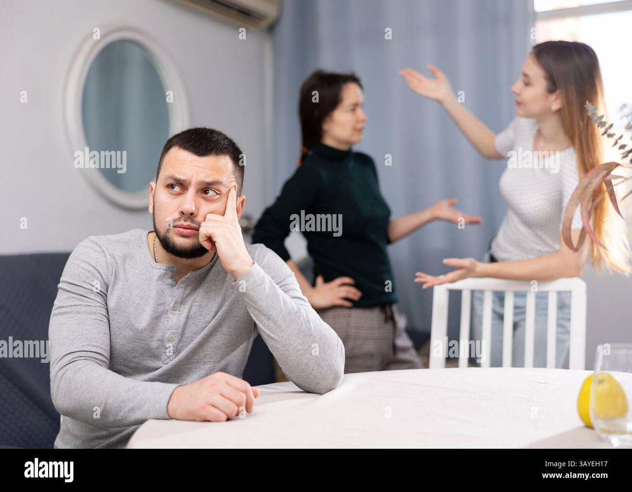 Triste uomo seduto a tavola a casa mentre due donne in lite Foto Stock