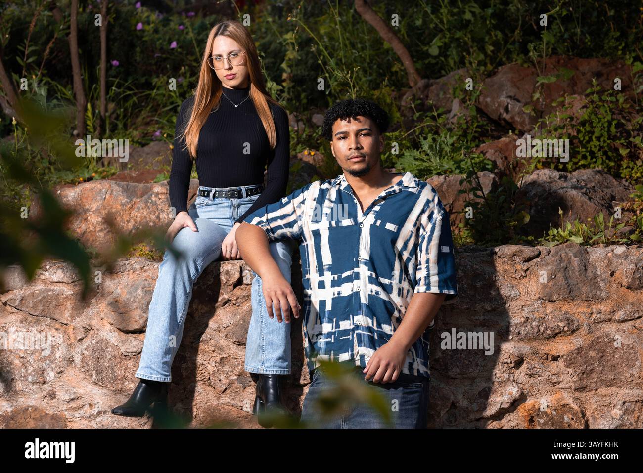 Amici alla moda che posano contro un muro di pietra rustico in un lussureggiante ambiente all'aperto Foto Stock