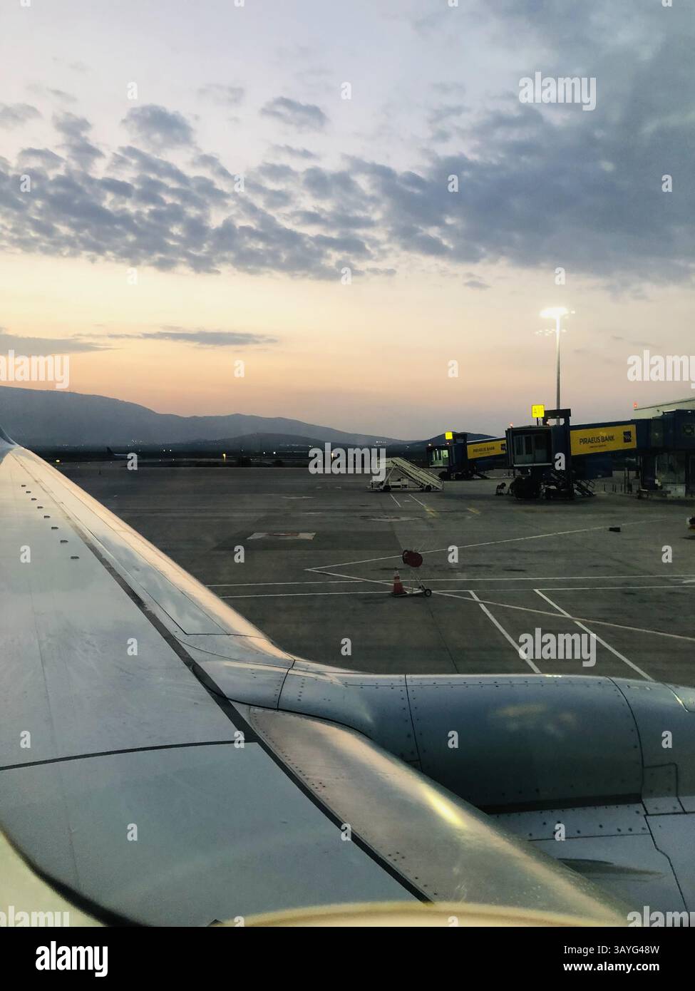 Una tranquilla vista del tramonto da un aereo di Transavia all'Aeroporto Internazionale di Atene, che mostra l'ala, il ponte jet con il logo della Banca del Pireo e la montagna distante - Immagine stock catturata con smartphone
