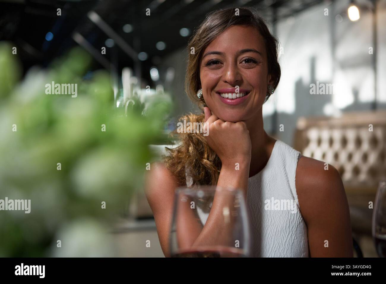 Donna che riposa il mento a portata di mano sorridente alla macchina fotografica del ristorante con bicchiere di vino e pezzo centrale verde Foto Stock