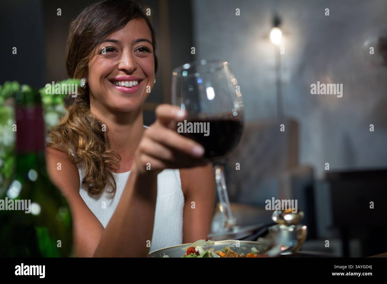 Donna ventenne che alleva un bicchiere di vino rosso al tavolo del ristorante, con bottiglia di vino, spazio per copiare Foto Stock