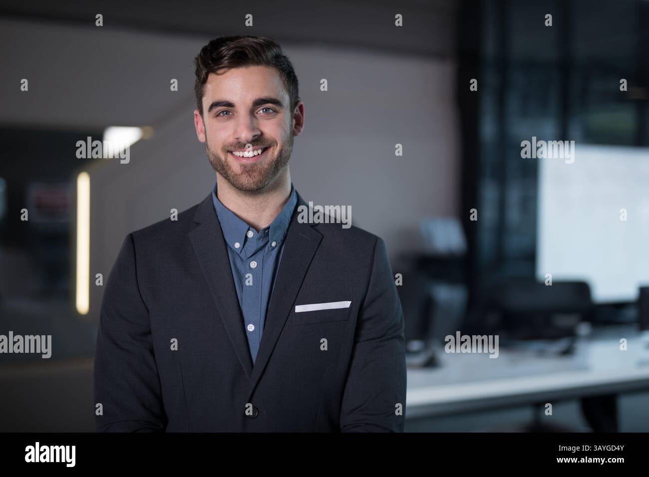 Giovane uomo adulto in piedi e sorridente in un ufficio open space, che espone tuta giacca e monitor Foto Stock